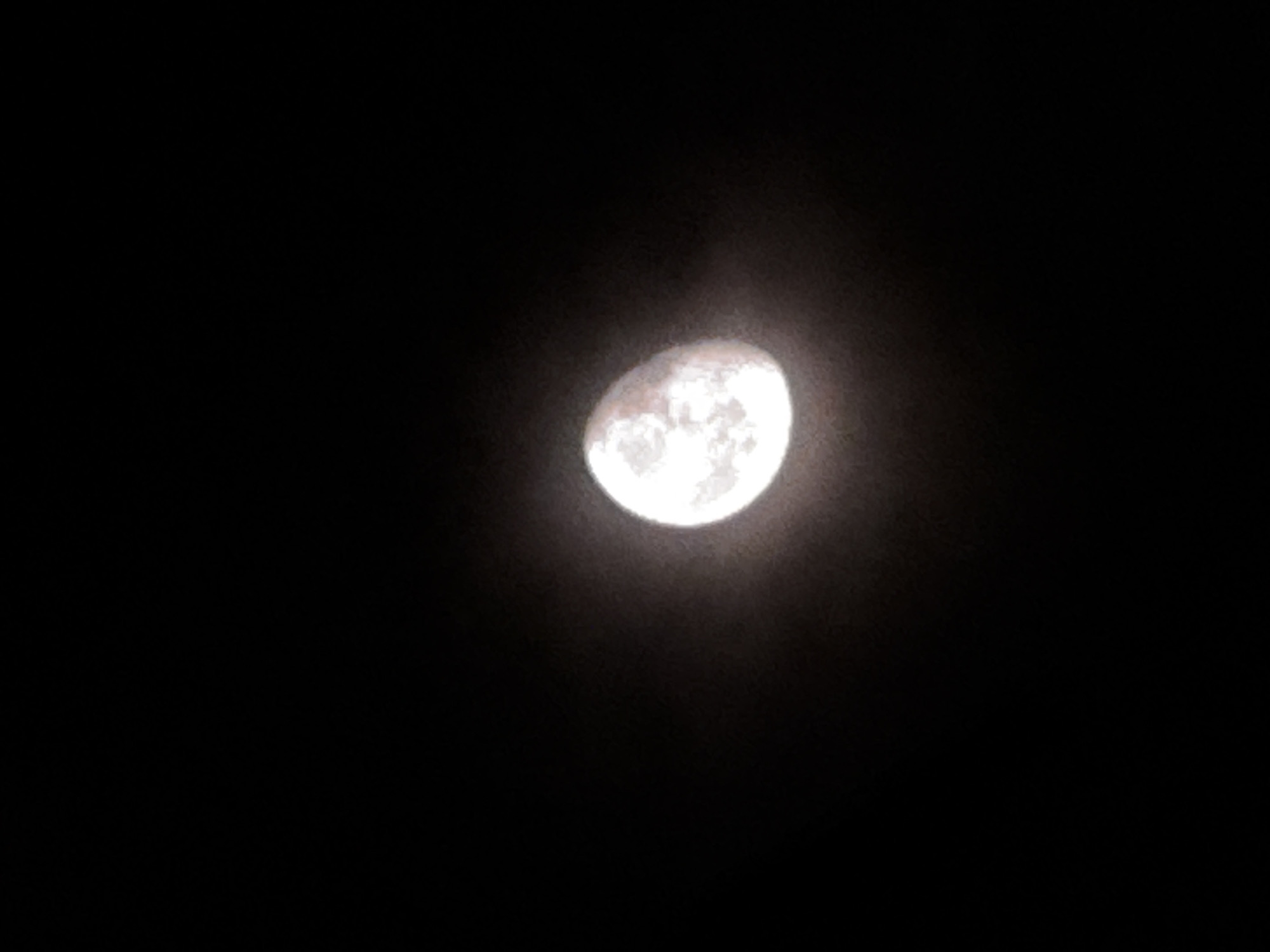 The image captures a brightly illuminated Moon set against a deep, dark night sky. The Moon is the central subject, appearing in a gibbous phase, with more than half of its surface visibly lit but not fully rounded. Its surface features are discernible, showing a mosaic of lighter and darker areas, characteristic of lunar topography.
A soft, hazy glow emanates from and surrounds the Moon, creating a gentle halo effect that diffuses into the blackness of the sky. The sky itself is completely devoid of stars, clouds, or any other visual elements, emphasizing the Moon's singular presence. The primary colors in the image are the luminous whites and greys of the Moon against the stark, profound black of the cosmos.
The scene unequivocally depicts nighttime. There are no discernible geographical features, man-made structures, or specific environmental cues within the photograph that would independently identify a particular location like Sofia, Bulgaria. The image focuses solely on the celestial body. No text is visible within the frame.