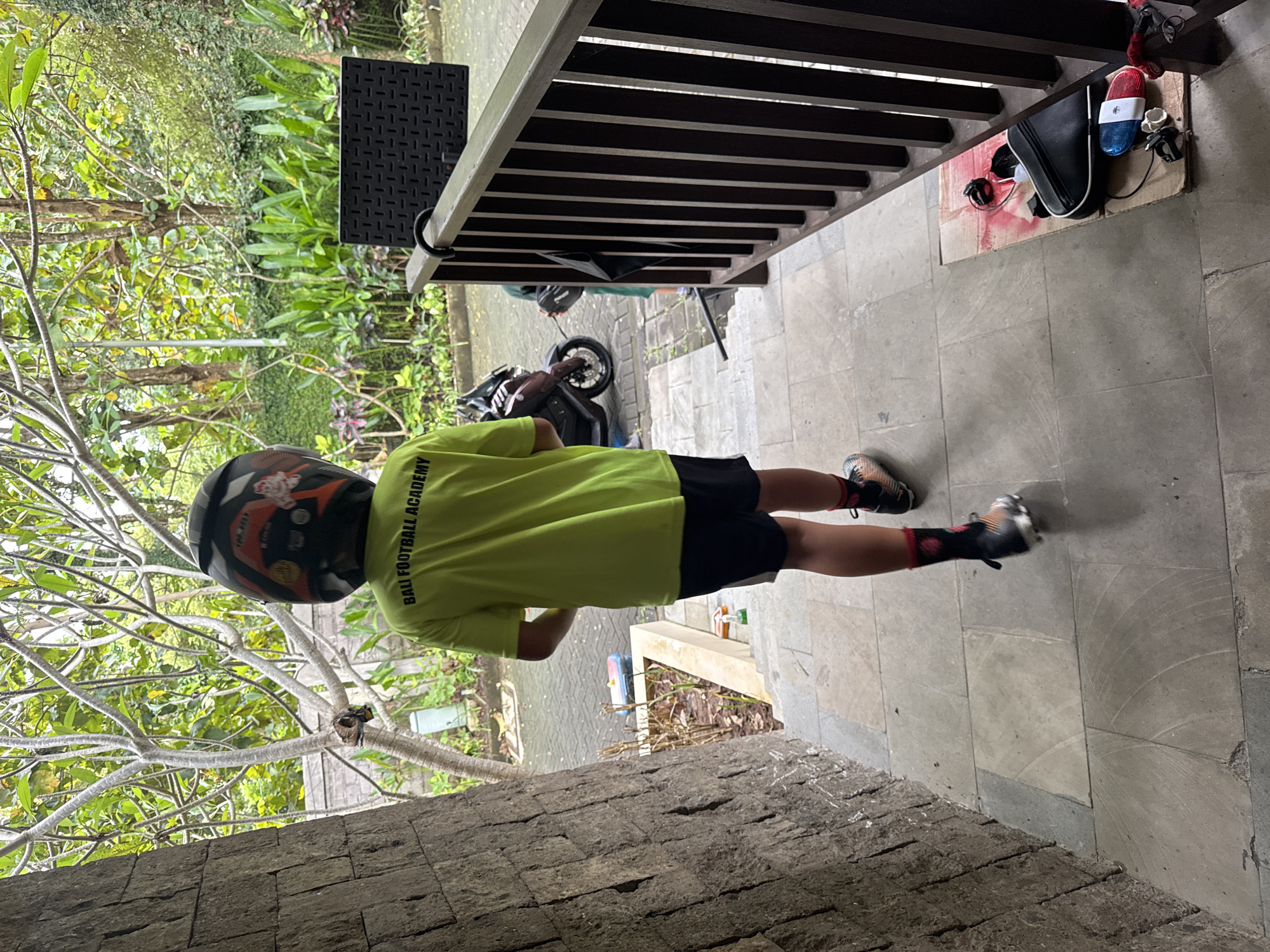 The image captures an outdoor scene, likely a patio or veranda, overlooking a lush tropical garden in Kuta Selatan, Indonesia. The ground is paved with large, light-grey square tiles, and a textured stone wall forms the foreground on the left.
The central subject is a young individual, seen from behind, wearing a black helmet with orange, red, and white design elements. They are dressed in a bright lime green T-shirt, black shorts, and athletic shoes with multi-colored patterns, paired with dark socks. The back of the T-shirt clearly displays the text "BALI FOOTBALL ACADEMY."
To the right, a dark wooden slatted railing extends horizontally across the frame. Beneath the railing, on the tiled floor, a black bag, a distinctive red and blue object resembling a flashing light or siren, and some loose wires are placed on a piece of cardboard. Further into the background, beyond the immediate patio, a black scooter or motorcycle is parked on a paved path.
The surrounding environment is rich with vegetation; mature trees with intricate, bare branches are visible in the left foreground, while a dense array of green foliage and plants fills the midground and background, characteristic of a tropical setting. A small, dark, square-patterned table or stool is partially visible on the far left. The scene is illuminated by natural daylight, suggesting either morning or late afternoon, and the overall atmosphere is calm. The person appears to be paused, possibly before or after a journey, given the helmet and scooter.