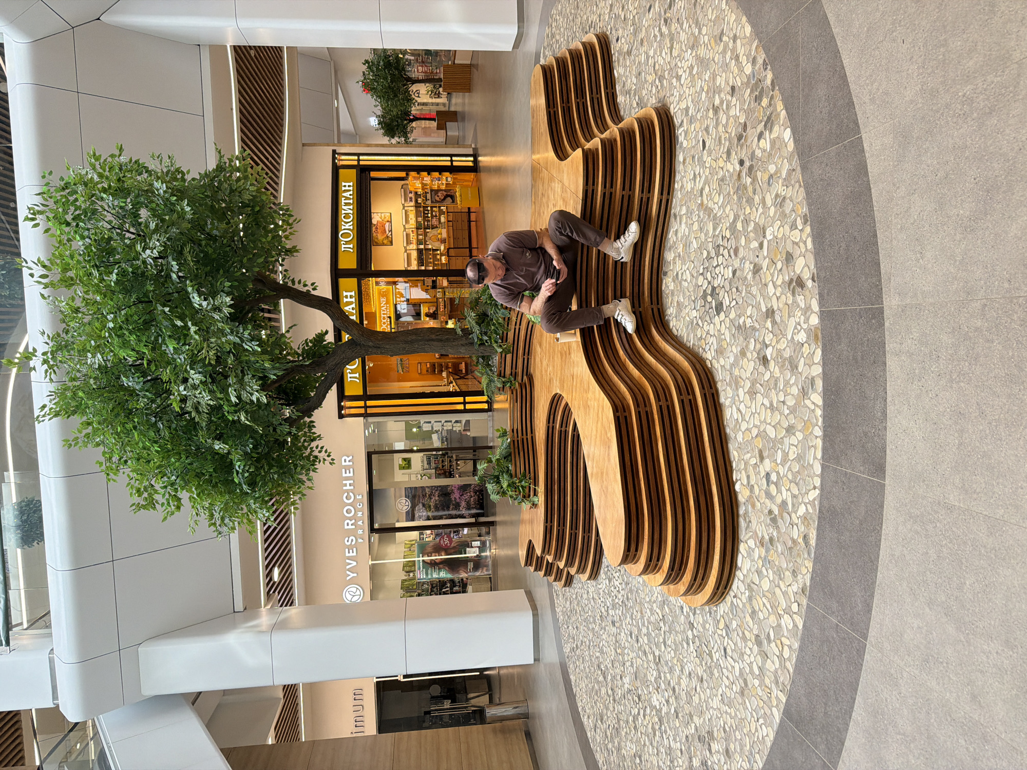 A man sits on a wooden bench in a shopping mall in Izhevsk, Russia. The bench is made of multiple layers of curved wood and is situated in an area with a pebble mosaic floor. A tree grows next to the bench, adding a natural element to the indoor setting. In the background, several stores are visible, including Yves Rocher and L'Occitane. The scene is well-lit and appears to be taken during the day.