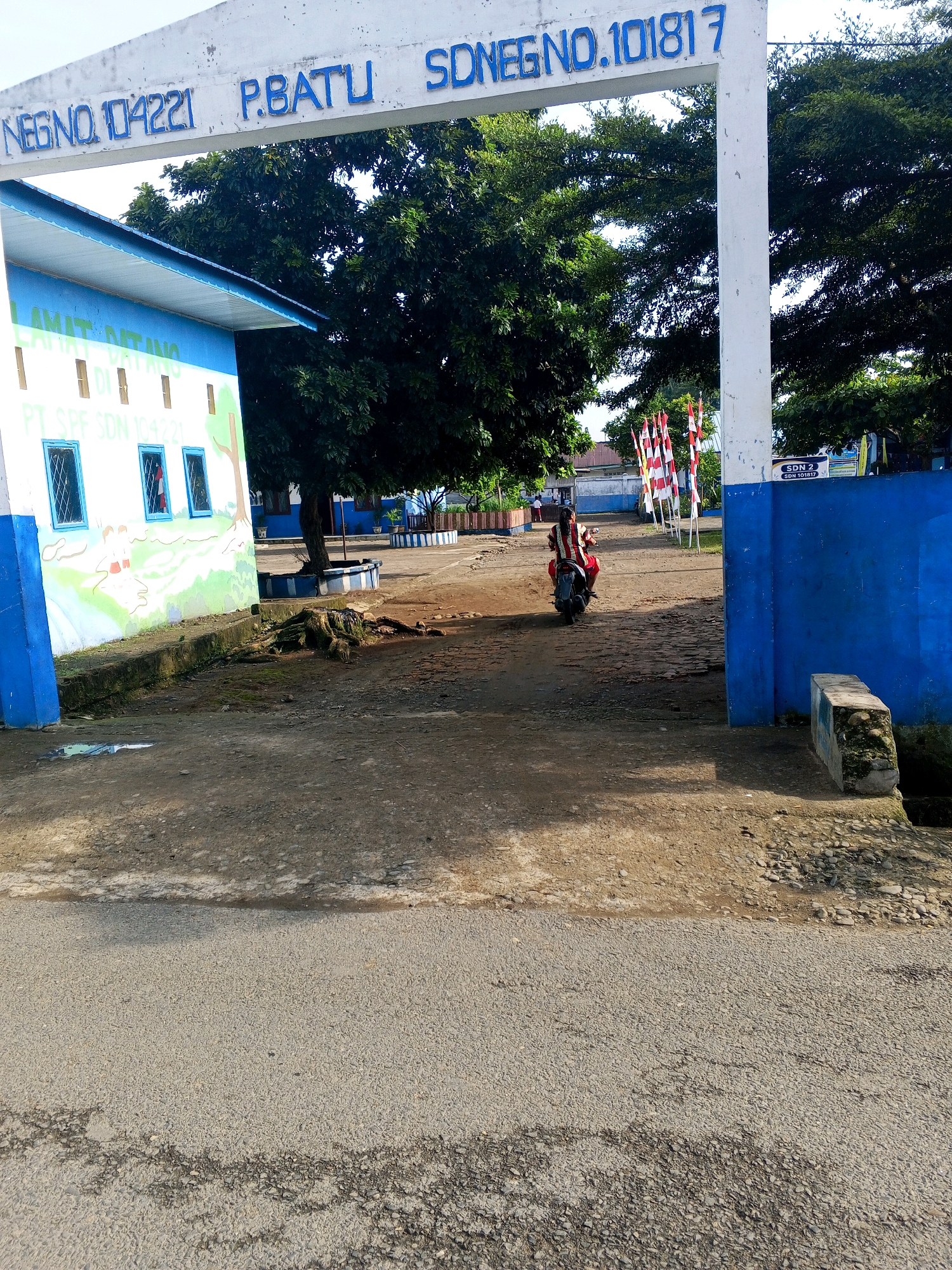 The image shows the entrance to a school named "SDN 2 P.BATU" in Pertampilen, Indonesia. The school's name is written in large blue letters on a white archway above the entrance. To the left of the archway is a white building with blue trim, featuring windows and a mural on the wall. A person is riding a motorcycle away from the entrance on a dirt path. The ground is a mix of dirt and asphalt. Several red and white flags are planted near the entrance. The overall scene depicts the exterior of a school building on a sunny day.
