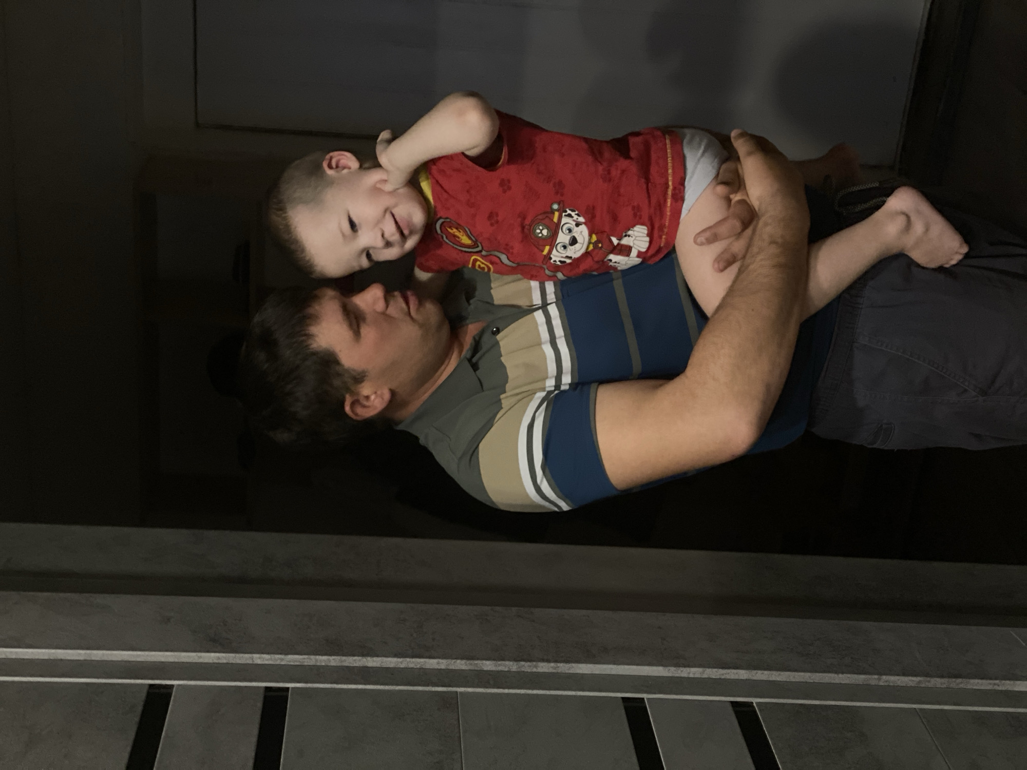 This image is a heartwarming indoor portrait featuring an adult male and a young child, captured in what appears to be a moment of affectionate interaction. The photo is rotated 90 degrees clockwise, so the subjects are oriented horizontally within the frame.
The adult male, likely a father figure, has dark hair and a medium build. He is wearing a multi-striped polo shirt with broad horizontal bands of blue, beige, and white, and dark-colored shorts or pants. He is holding the child securely in his arms, cradling the child's back and supporting their legs. His gaze is directed towards the child, and his expression appears gentle and engaged.
The young child, a toddler, has light hair and fair skin. They are wearing a red t-shirt featuring the character Marshall from the "Paw Patrol" animated series, and light-colored diaper or underwear. The child is positioned face-to-face with the adult, lying on the adult's chest or shoulder area, and is looking directly at the adult. A wide, joyful smile is spread across the child's face, indicating happiness and engagement in the interaction. One of the child's hands is near their ear, possibly scratching or just in motion during play.
The setting appears to be dimly lit indoors, with the background being mostly dark and indistinct, suggesting a room or hallway with low lighting. At the bottom of the original vertical frame (which is the left side of the current rotated image), there's a visible floor or step area composed of grey tiles or panels with horizontal lines and darker gaps, adding some texture to the foreground.
The overall mood of the image is tender and loving, capturing a special bond between the two individuals. There is no discernible text other than the character on the child's shirt.