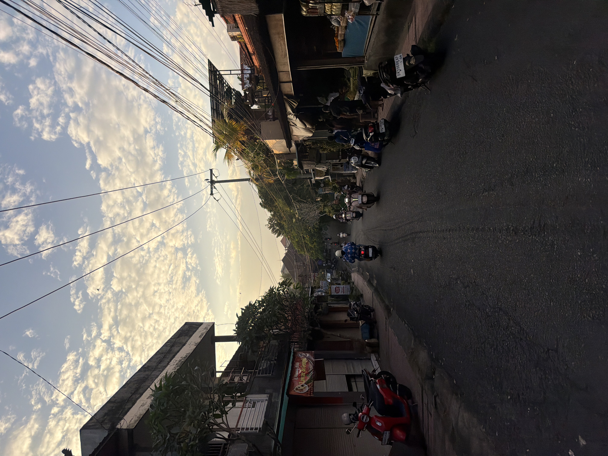 The image depicts a street scene in Ubud District, Indonesia, at what appears to be either sunrise or sunset, indicated by the warm light and scattered clouds in the sky. The street is paved and slopes gently upwards, lined with buildings on both sides. On the left, a multi-story building with dark walls and balconies is visible, with a red scooter parked on the sidewalk in front of it. Several shops with closed shutters are also present on this side.
On the right side of the street, there are more shops, some with awnings and displays of goods. People are seen standing and walking in front of these establishments. Numerous motorbikes are present, either parked along the street or being ridden. Several individuals are riding motorbikes, some in the foreground and others further down the street, suggesting typical street activity. The environment includes trees, power lines strung across the street, and various architectural styles of the buildings, characteristic of a Southeast Asian urban or semi-urban setting. The overall impression is one of a busy street in a town or city, with motorbikes being the primary mode of transportation.