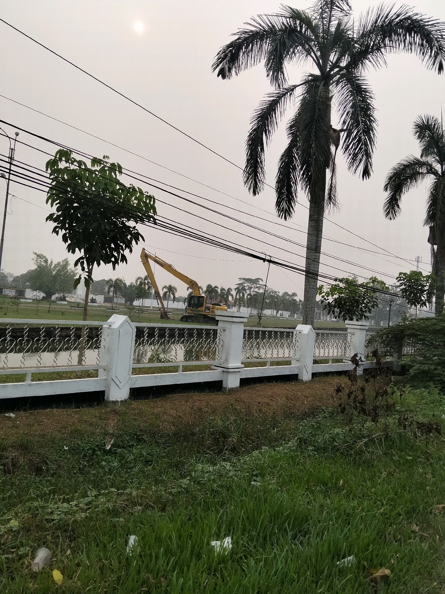 The image captures an outdoor scene in Sungai Raya, Indonesia, characterized by an overcast sky and a pale sun. In the foreground, a grassy area with scattered litter leads to a white decorative fence. Beyond the fence is a body of water, likely a canal or pond, with a reflection visible on its surface. In the middle ground, a yellow excavator is positioned near the water's edge, suggesting ongoing construction or maintenance work. Several palm trees and other greenery dot the landscape, with power lines stretching across the upper portion of the frame. The background shows more palm trees and buildings, possibly part of a residential or park area. The overall impression is one of a somewhat neglected semi-urban environment undergoing some form of development.