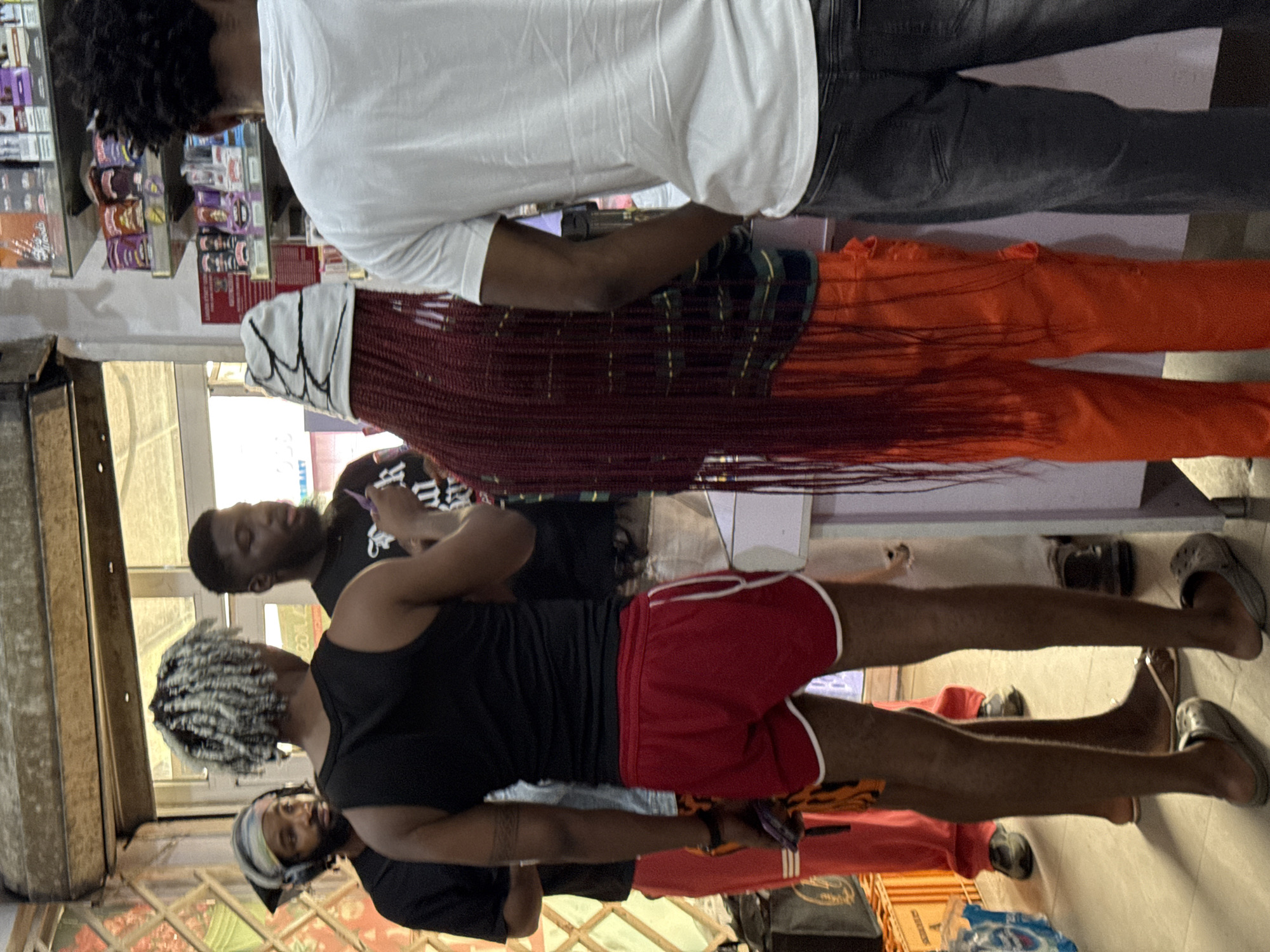 The image shows a street scene in Ado, Nigeria. Three men are standing in front of a shop. One man wears a white t-shirt and grey jeans. Another man is wearing orange pants. A second man is cutting the hair of a man wearing red shorts. A third man is standing behind the man in red shorts. The background features shop merchandise. The image is rotated clockwise.