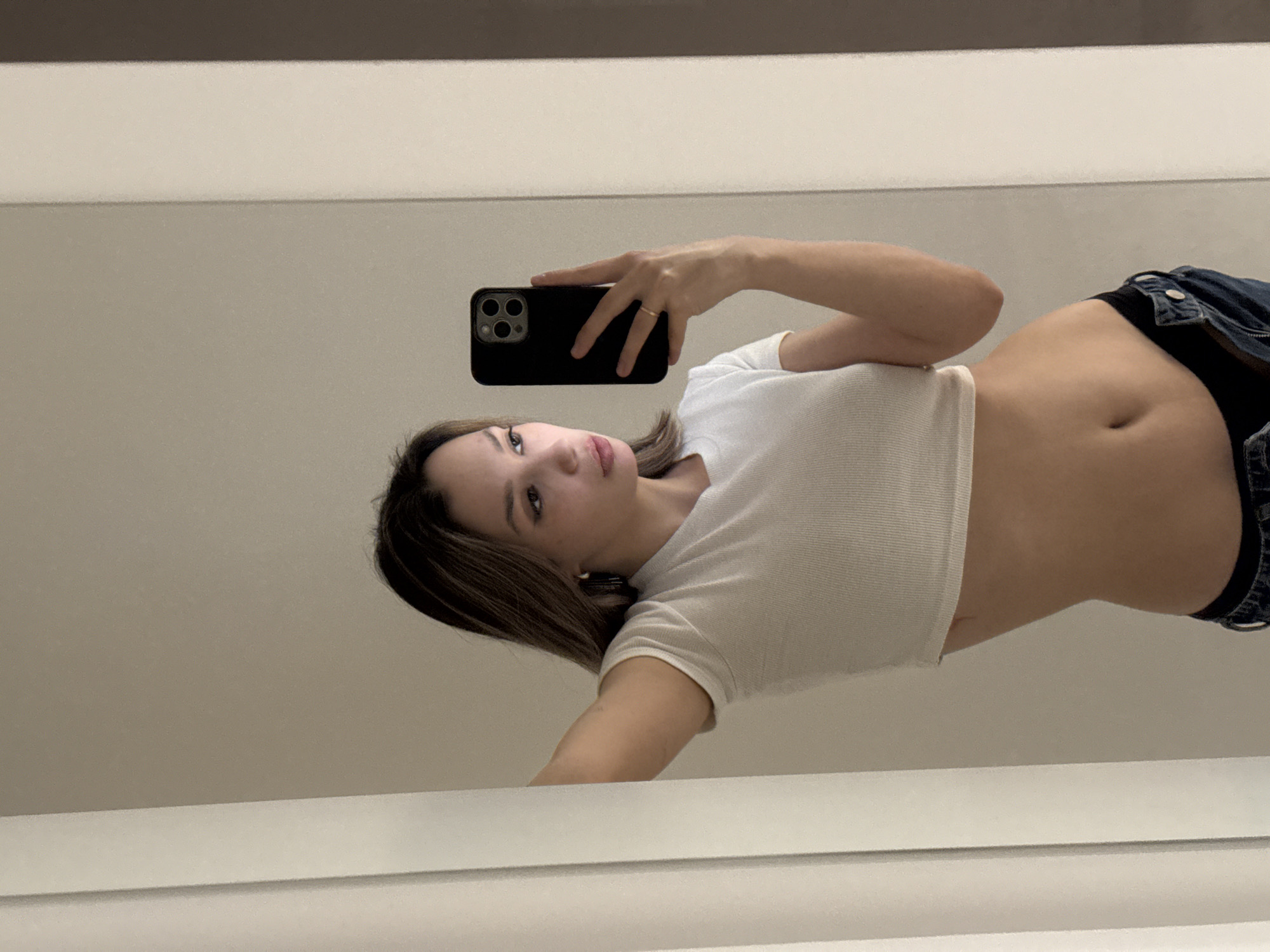 A young woman is captured in a mirror selfie, lying or leaning in a way that shows her torso angled upwards from a low perspective. She holds a black smartphone with a triple-lens camera in her right hand, aiming it towards herself. She wears a simple gold-colored ring on her right ring finger. Her head is tilted, and she looks directly towards the camera with a neutral expression and slightly parted lips. Her hair is medium length and brown, parted to the side.
She is dressed in a white, waffle-knit style cropped long-sleeve top, revealing her midriff and navel. Below the crop top, she wears black underwear or bikini bottoms, and unzipped blue jeans are visible around her hips.
The background, which is the reflection of the room she is in, consists mainly of a plain, light-colored surface, possibly a wall or ceiling, with a darker horizontal stripe running across the top portion of the image. The lighting appears even and bright. There is no visible text in the image.