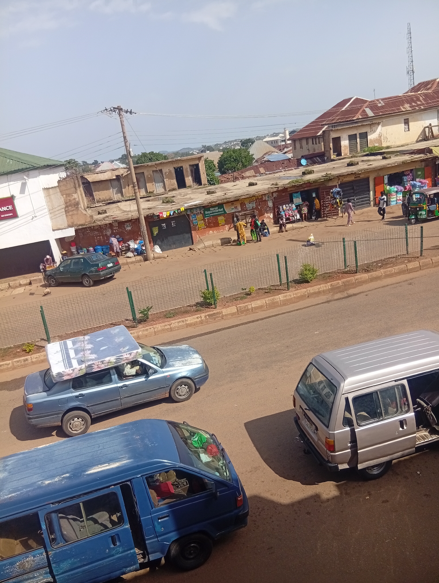 In Bukuru, Nigeria, a street scene unfolds with several vehicles and pedestrians. A blue van with its doors open is parked on the right side of the road. A light blue sedan is traveling in the foreground, carrying a mattress on its roof. Further down the road, a dark green sedan is parked. In the background, a green and white tricycle is visible, along with several people walking and standing near storefronts. The buildings appear to be commercial structures, some with corrugated metal roofs. The sky is clear and the ground is paved.