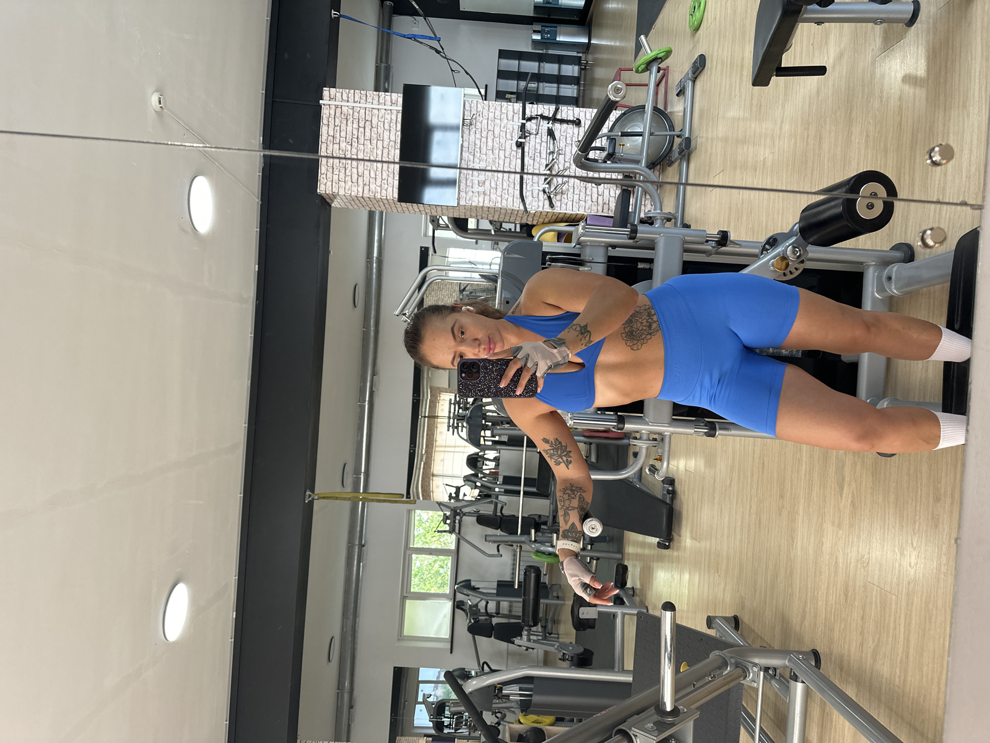 The image depicts a fitness center in Yuzhnouralsk, Russia, observed through a large, reflective mirror. In the foreground, a woman is captured in a self-portrait, holding a dark-colored smartphone with a patterned case in her right hand, positioned to capture her reflection. She is an adult with dark hair pulled back, wearing a bright blue athletic two-piece outfit consisting of a sports bra and high-waisted shorts, along with white ankle socks and white fingerless lifting gloves. White wireless earbuds are visible in her ears, and a light-colored wristband is on her left wrist. She has several visible tattoos, including floral designs on her left arm and right torso.
The mirror reflects the spacious gym interior. The flooring is light-colored, resembling wood laminate. The walls are predominantly white, with a dark grey horizontal band running across the upper section. The ceiling is white, featuring circular recessed LED lights and faint linear marks, possibly from previous water exposure. A white smoke detector is visible on the ceiling in the upper right.
Numerous pieces of silver-framed gym equipment with black upholstery are scattered throughout the space. On the left, a leg press machine is prominent. In the mid-ground, a brick-veneered pillar stands, with a flat-screen television mounted on its side. A yellow strap hangs from the ceiling near this pillar. Further back, rectangular windows reveal green foliage outside, indicating a daytime setting. Additional gym machinery, including benches, weight plates (some green), and a water cooler, are visible in the background. A vertical seam or edge of the mirror panel runs down the right side of the image.