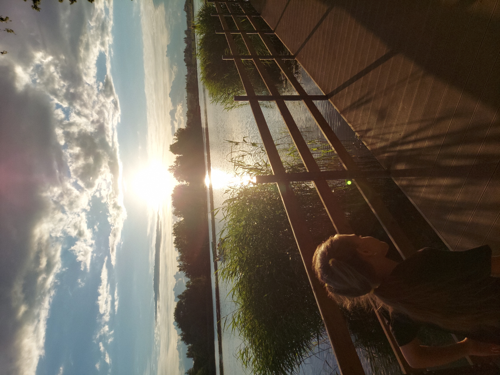 A person is sitting on a wooden boardwalk overlooking a body of water. The sun is low in the sky, casting long shadows and a warm glow across the scene, indicating it is either sunrise or sunset. The water is calm, with tall reeds growing along the bank. In the distance, there is a tree line and a hazy sky filled with clouds. The boardwalk has a rustic, wooden railing. The person is viewed from the side and appears to be looking out at the water and sky. The overall mood is peaceful and serene. The image was taken in Razvetye, Russia.
