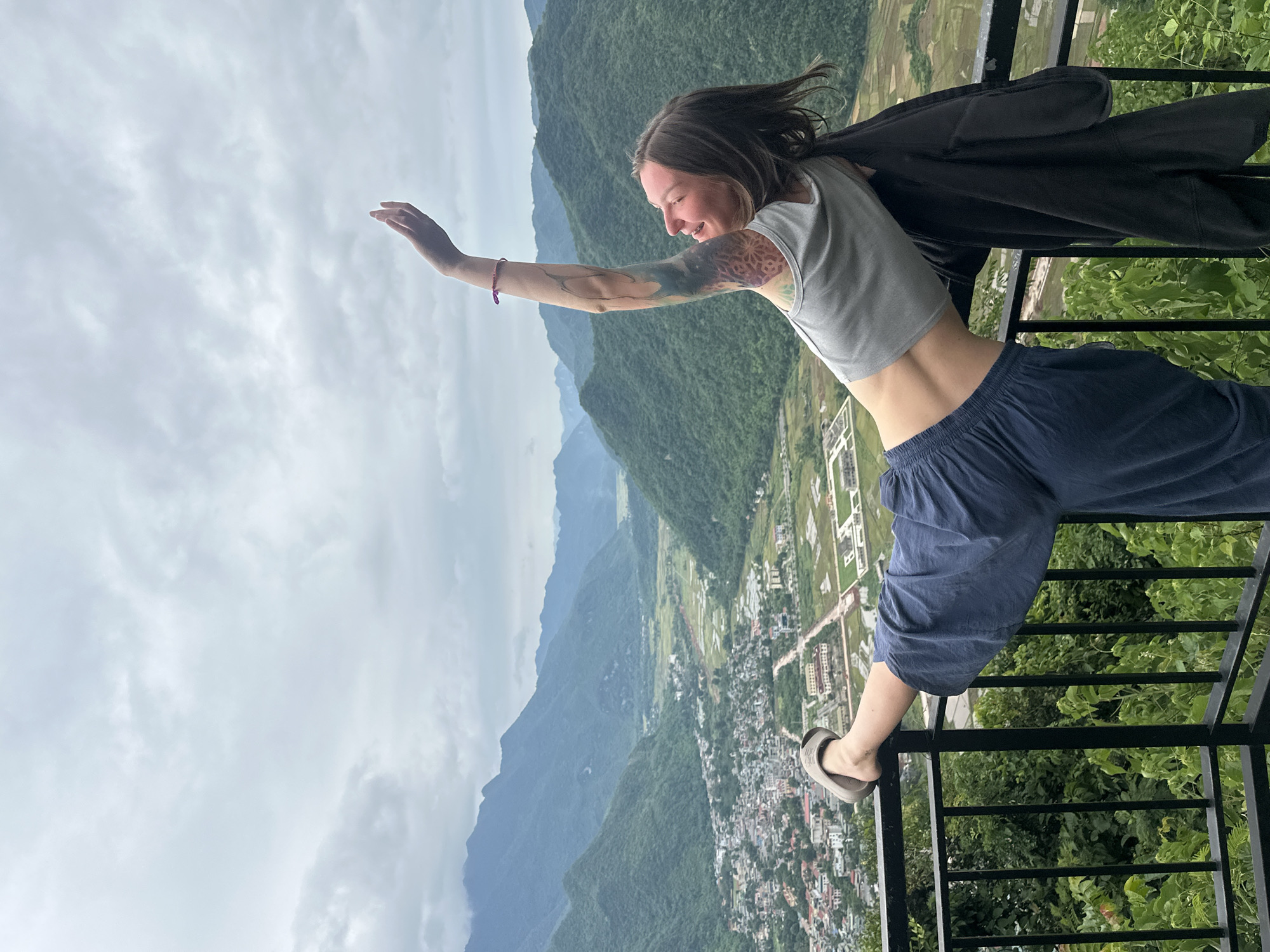 A young woman with tattoos on her left arm leans over a black metal railing overlooking a valley in Mai Chau, Vietnam. She wears a grey crop top and blue pants, with one leg extended and resting on the railing. Her right arm is raised above her head, and she is smiling. The background features lush green mountains and a town nestled in the valley below. The sky is overcast with clouds.