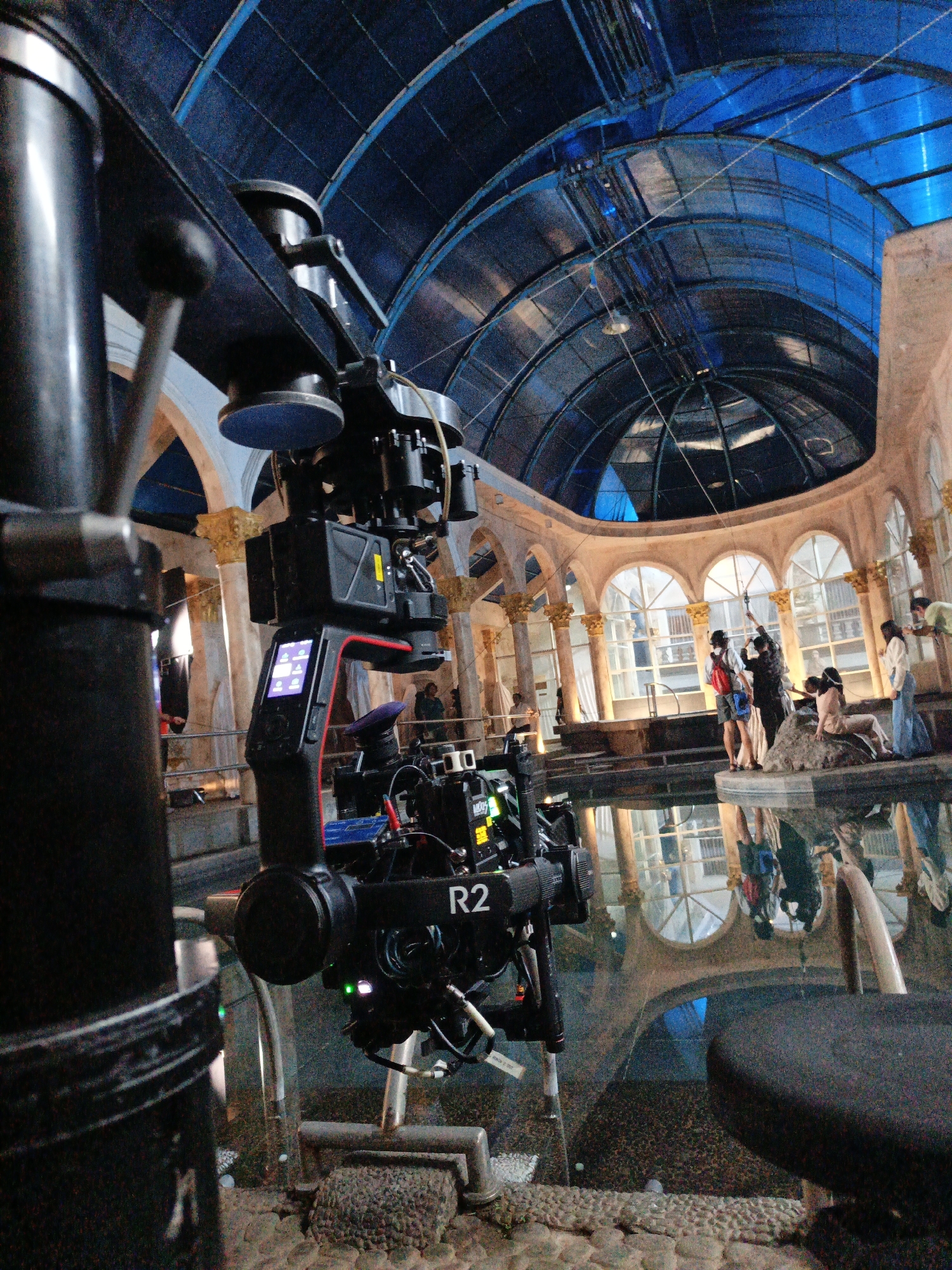 A scene is captured within a covered atrium, likely a film set in Cibogo, Indonesia. The structure features a domed, blue translucent roof and arched windows. In the foreground, a camera rig with a prominent "R2" label is positioned. The rig is stabilized and appears to be in operation, with a screen displaying icons. In the mid-ground, several individuals are gathered around a central stone element, with some posed as if in a tableau. The area appears to be partially filled with water, reflecting the architectural elements and the sky. The overall impression is that of a staged environment for a production.