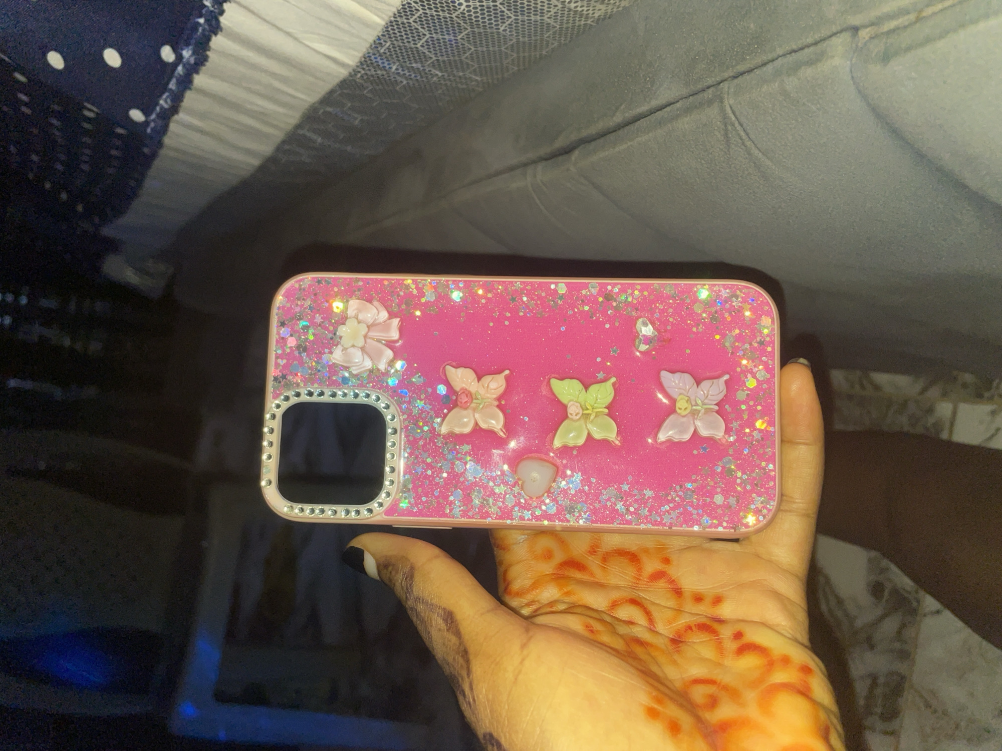 A person's hand, adorned with henna, is holding a pink smartphone case decorated with glitter, rhinestones, and small butterfly and bow embellishments. The case is placed on a dark, textured surface. In the background, a white and navy blue patterned fabric is visible, along with a shadowy, dimly lit environment. This image was taken in Keffi, Nigeria.