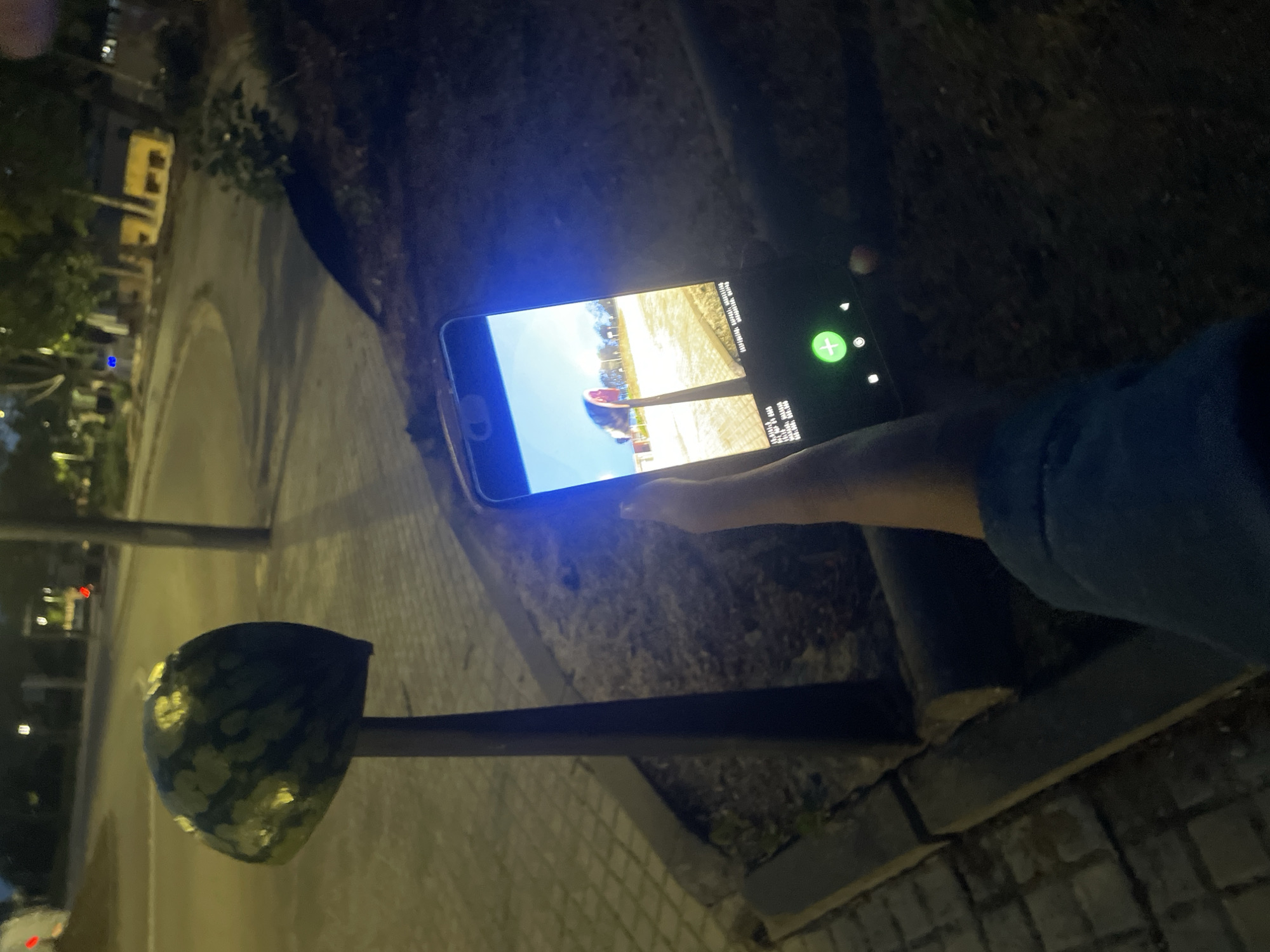 The image, rotated counter-clockwise, depicts a night scene outdoors, likely on a street or sidewalk in La Granada, Spain. A person's right arm and hand are visible on the right side, holding a black smartphone horizontally. The hand has a light skin tone, and the arm wears a dark blue long-sleeved garment.
The smartphone screen is brightly illuminated, displaying an application. The screen shows a vibrant outdoor scene under a clear blue sky, featuring a light-colored ground or path, trees or foliage in the distance, and a prominent red, possibly curved, structure in the foreground of the displayed image. On the right side of the phone's screen, some small, light-colored text appears, likely displaying technical information or statistics such as "GPU", "CPU", and numeric values, consistent with a gaming or demanding application. A green circular button with a white plus sign is also visible on the right edge of the phone screen, suggesting an interactive element.
To the left of the hand and phone, a dark, slender pole rises from the ground, topped with a distinctive, rounded, dark object patterned with lighter, possibly reflective, green or yellow shapes. This object resembles a decorative street lamp or bollard. The ground beneath the pole and the person is paved with square or rectangular tiles. In the background, illuminated by distant artificial lights, are hints of buildings with visible windows and surrounding dark foliage. The overall lighting suggests it is nighttime.