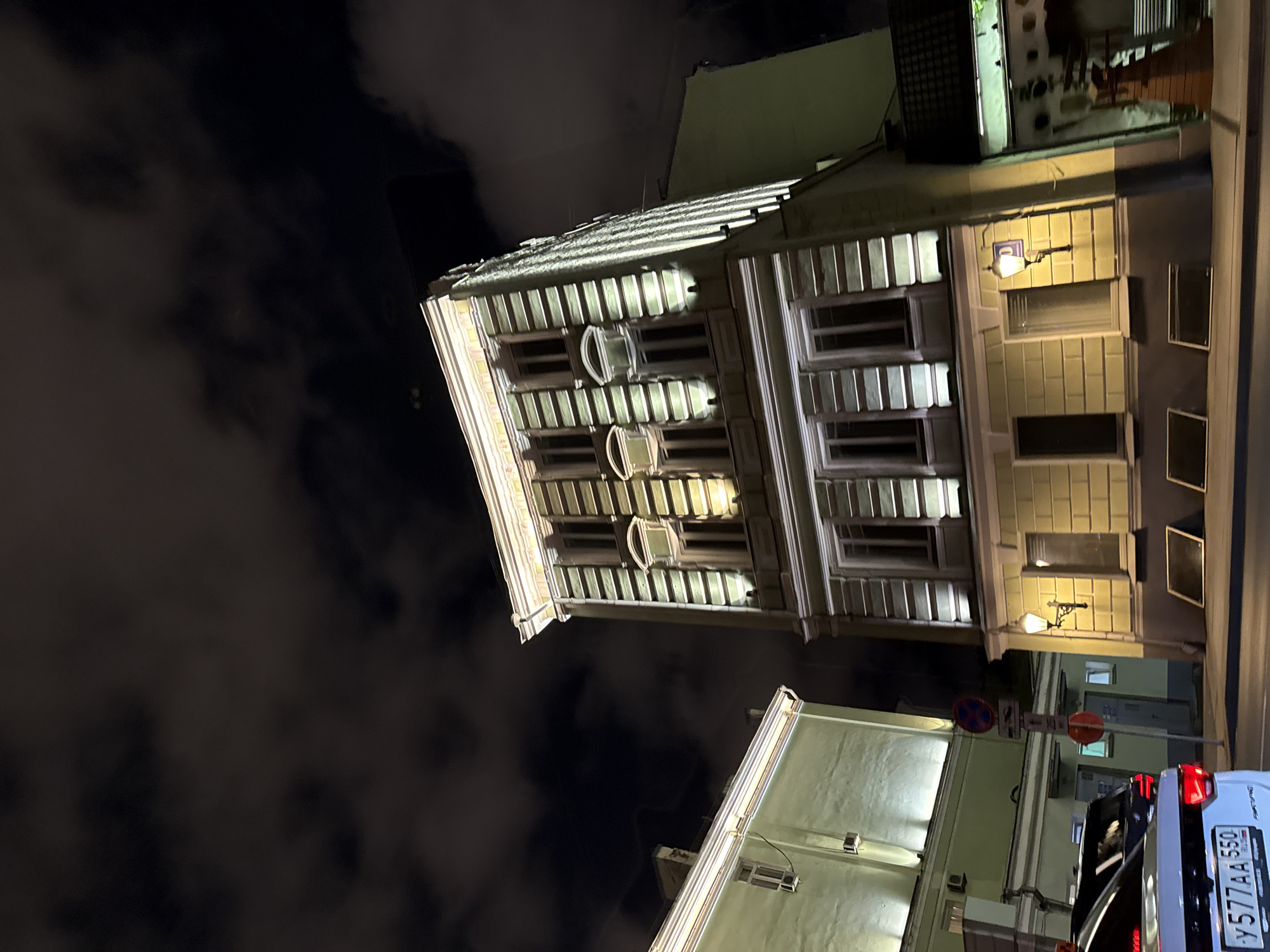 The image shows a street scene at night in Moscow, Russia. The focus is on a historic, pale yellow building with multiple windows and decorative architectural elements, illuminated by spotlights. To the left, another building, painted a light green, is partially visible. In the foreground, the rear of a white car with Russian license plates (Y 577AA 550) is parked. Other vehicles are also present on the street. Traffic signs are visible on a pole between the buildings. The sky is dark and cloudy, suggesting night time. The overall impression is a well-maintained urban environment at night.