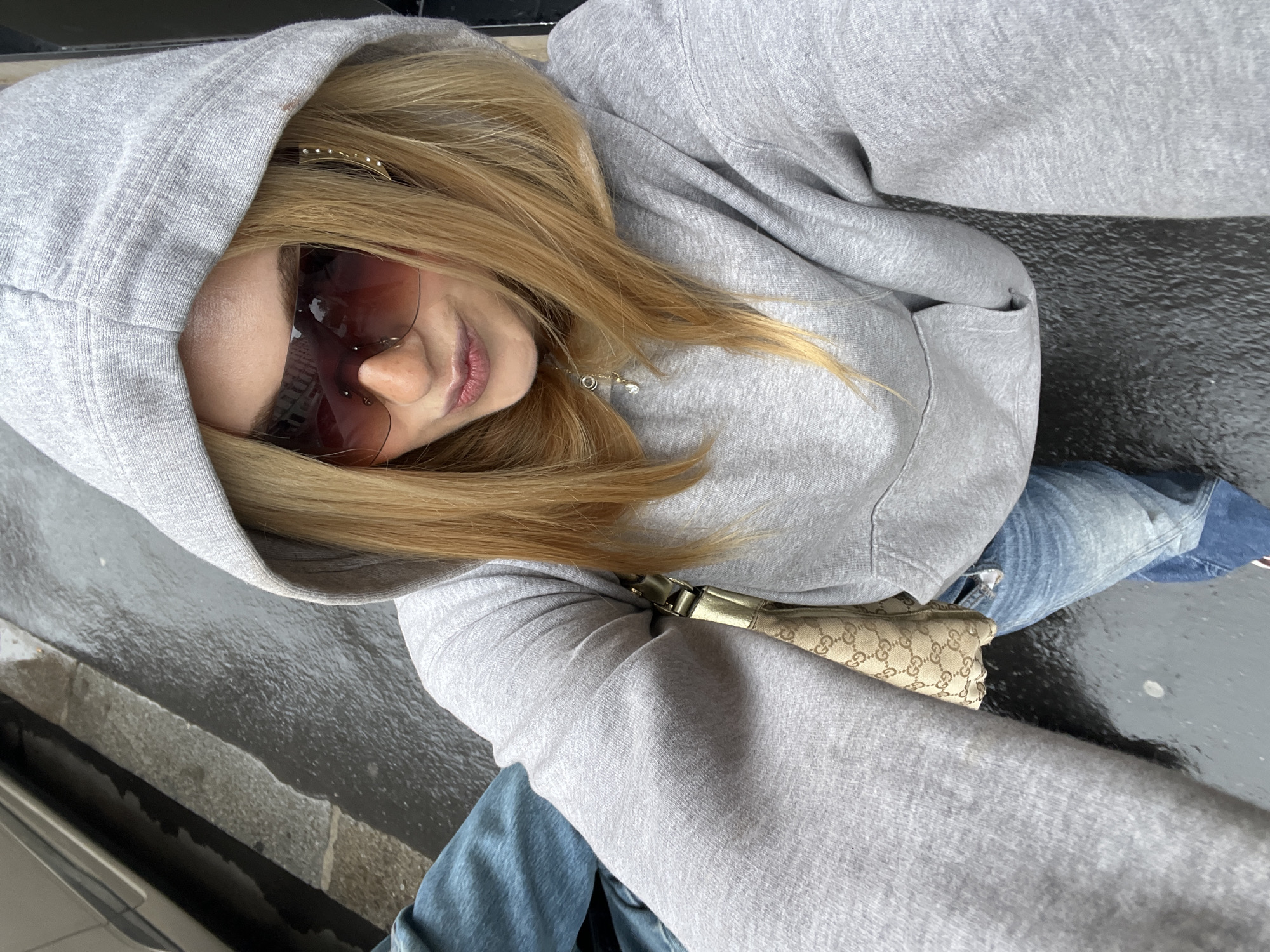 A woman wearing a grey hoodie, sunglasses, and jeans is taking a selfie in an urban environment. She is holding the camera with her right arm, positioned above her head, creating a downward angle of the shot. The background appears to be a street with dark, possibly wet pavement. A beige-colored bag with a pattern is visible across her torso. The overall setting is consistent with an outdoor location, possibly in a European city, based on the architecture and street features. There is no explicit information in the image to confirm it is Paris.