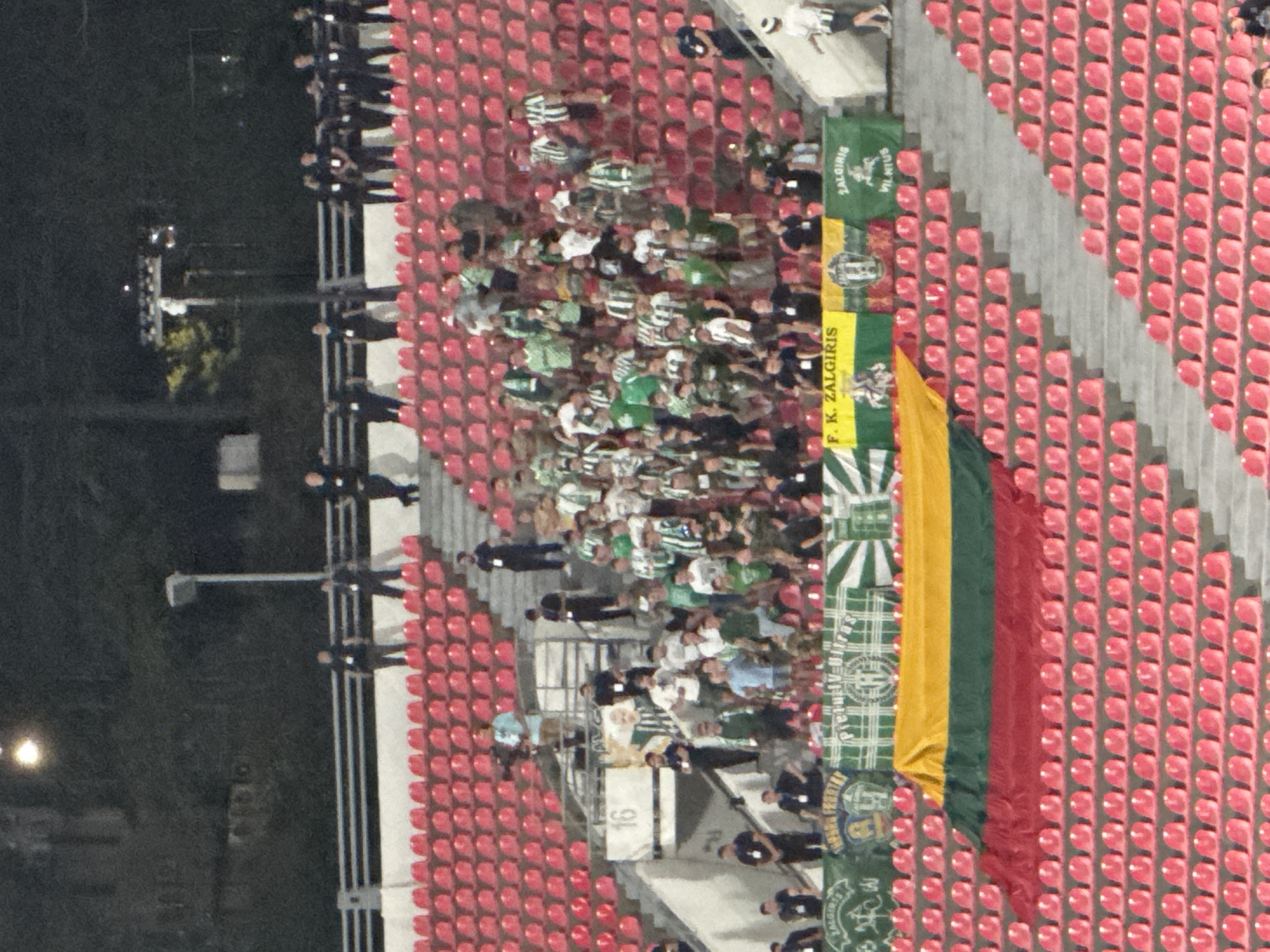 An aerial shot captures a stadium with tiered red seating filled with spectators watching an event. In the foreground, a large Lithuanian flag, composed of yellow, green, and red horizontal stripes, is draped across some of the seats. Behind the flag, several banners are displayed, some featuring green and white stripes and text. A group of people wearing green and white striped jerseys are visible in the stands, suggesting they are supporters of a sports team. There are also individuals in dark uniforms, possibly security personnel or staff, positioned throughout the seating areas and along the walkways. The background reveals more stadium architecture and distant trees, indicating an outdoor venue. The scene appears to be taking place at night or in low-light conditions, with artificial lights illuminating parts of the stadium.