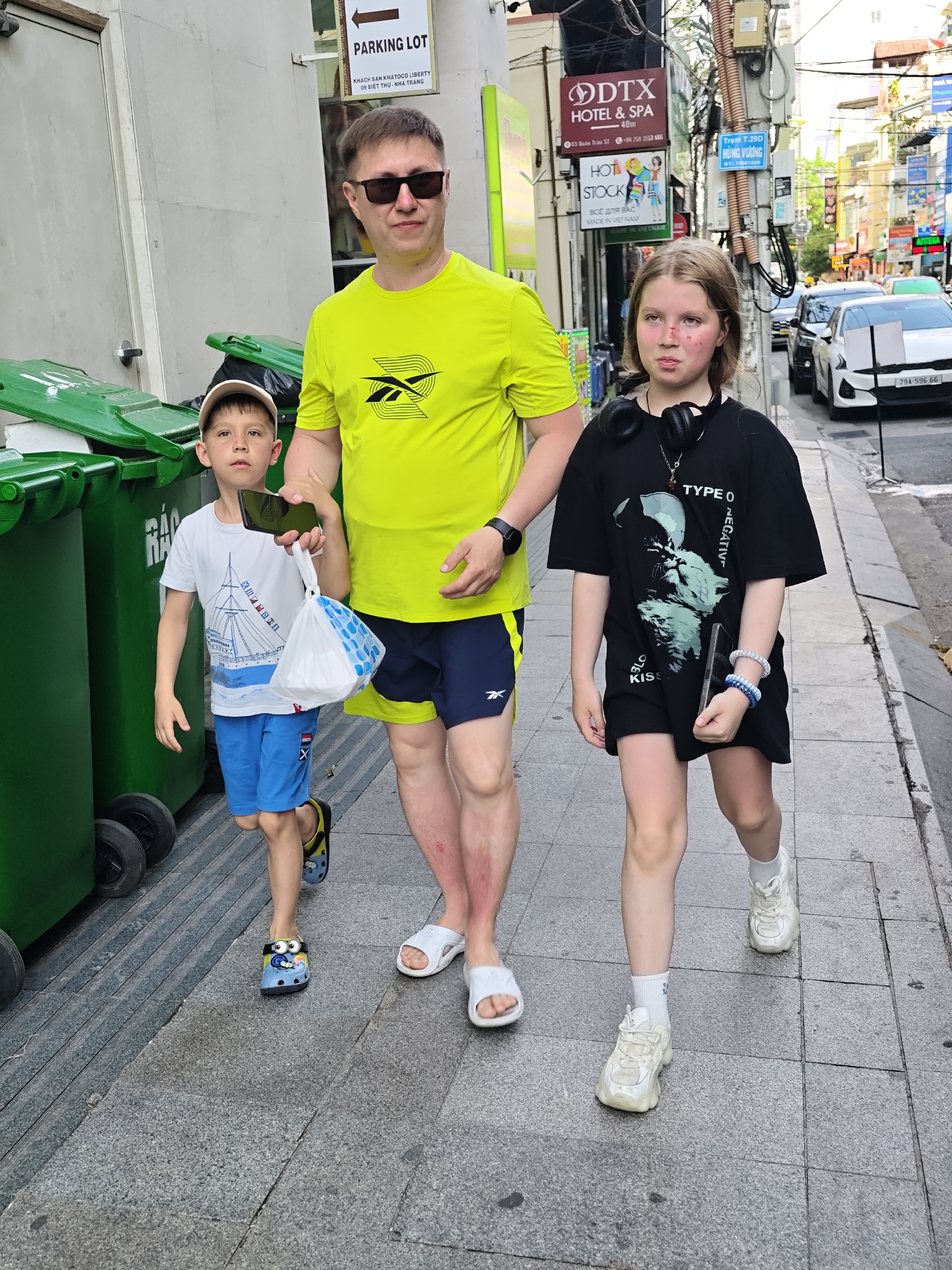 In Nha Trang Ward, Vietnam, on a sunny day, a man, a young girl, and a young boy walk along a paved city sidewalk next to a row of green trash bins. The man, on the left, wears a bright yellow t-shirt and dark shorts. He holds a smartphone in his right hand. Beside him, the young girl, in the center, wears a black t-shirt with a cat graphic and a short skirt, with headphones around her neck. She carries a smartphone in her right hand and wears bracelets on her left wrist. The young boy, on the far left, is dressed in a white t-shirt and blue shorts and carries a plastic bag with a blue and white pattern and a smartphone. The sidewalk is made of grey tiles, and the background shows buildings with signs, including one for "DTX HOTEL & SPA". A white car is visible parked on the street in the background.