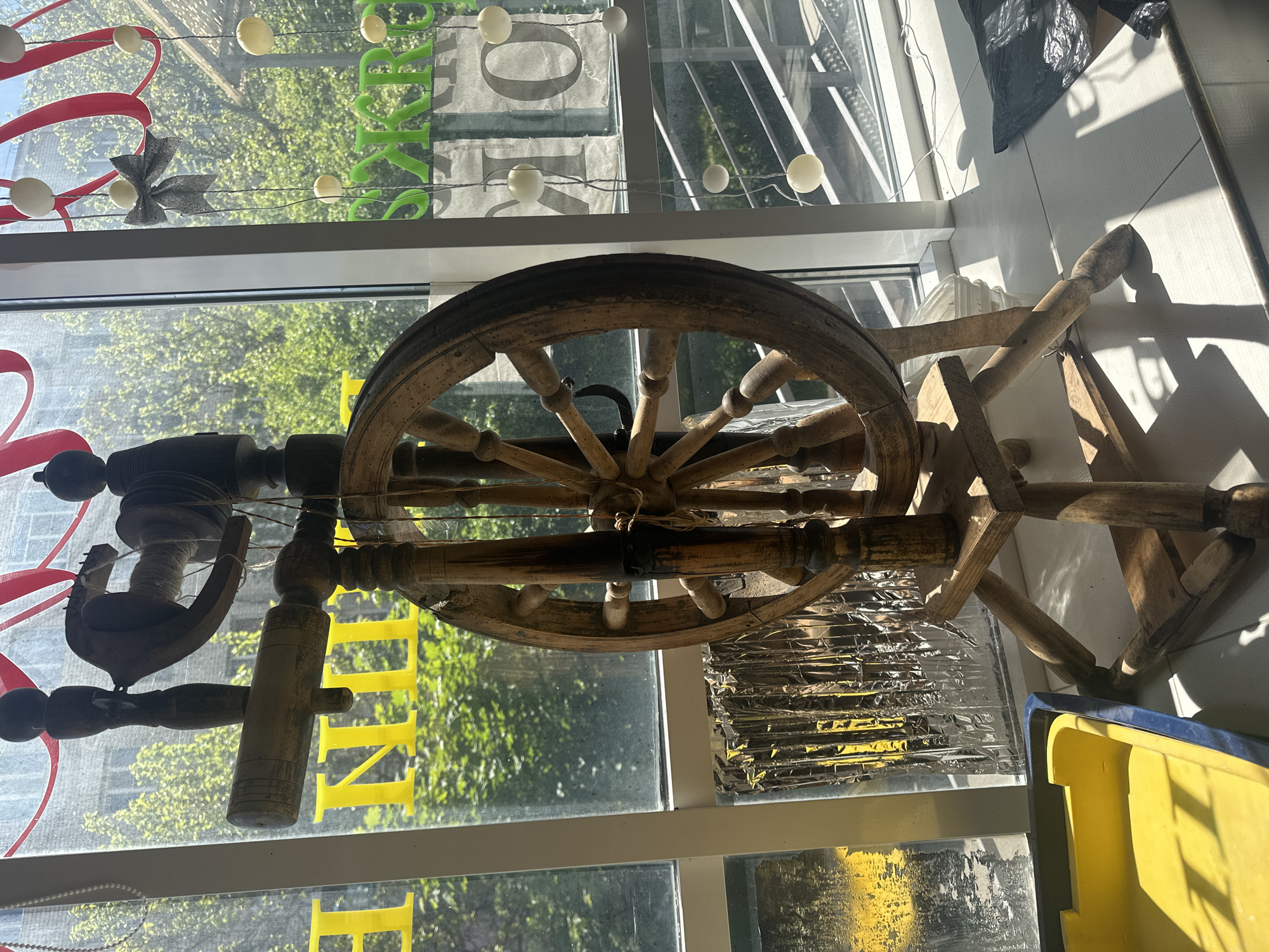 The image displays an indoor scene looking out through a large window on a bright, sunny day. A vintage, rustic wooden spinning wheel is centrally positioned, dominating the foreground. Its large wheel, spokes, and various turned wooden components are visible, showing signs of age and minor imperfections.
Through the window, the vibrant green foliage of trees is clearly visible, suggesting an outdoor natural setting. The sunlight casts strong, sharp shadows on the indoor surfaces, indicating a clear, sunny day.
Decorative elements are strung across the upper portion of the windowpane: a string of white, spherical globe lights, a flowing red ribbon-like decoration, and a grey glittery bow.
Faint text is visible through the window, likely from signs. Green letters "RY" can be seen towards the upper right, and partially obscured yellow letters, possibly "IN" and "E", are visible on the lower left.
To the right of the spinning wheel, on a tiled surface or ledge, sits a black plastic bag and a bright yellow plastic container or bin. Below the spinning wheel, there is a section of reflective, silver-colored material, possibly foil or insulation, with hints of yellow underneath. The arrangement suggests a display, potentially in a shop window or a creative workspace.