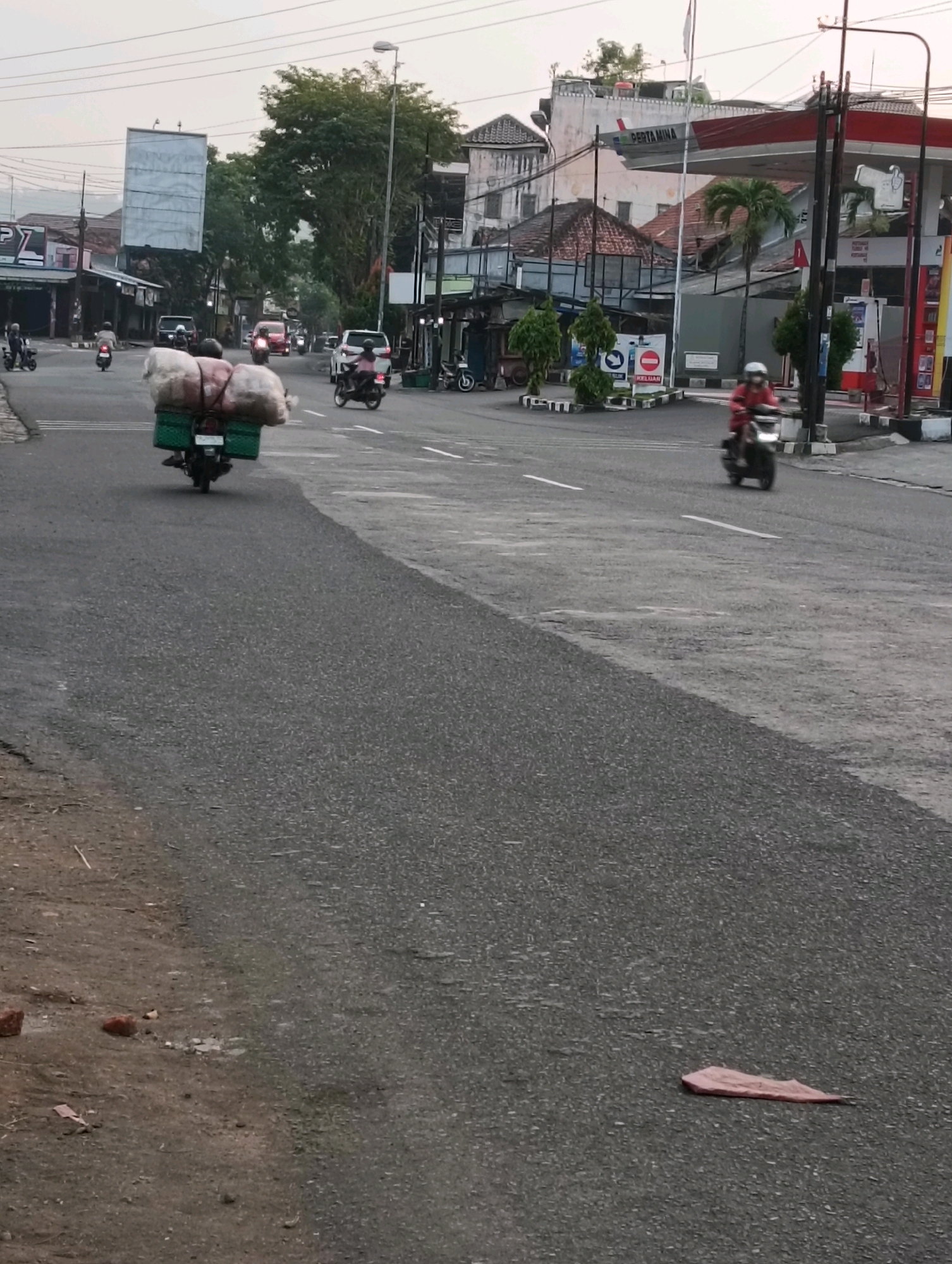 This is a street scene in Purworejo, Indonesia, likely during daylight hours, possibly early morning or late afternoon given the lighting. The image captures a road with traffic, including several motorcycles and cars. In the foreground, a motorcycle is being ridden by an individual wearing a helmet, carrying a large load of what appears to be bundled goods secured to the back of the motorcycle with green strapping. The goods are a light color, possibly fabric or sacks of produce. Further down the road, another motorcycle rider wearing a helmet and a red jacket is visible. On the right side of the road, a Pertamina gas station is prominent, with its distinctive red and white signage. Buildings lining the street appear to be commercial establishments, some with awnings. Trees are present along the roadside, and utility poles and wires are visible. The road surface is asphalt with visible lane markings. The overall environment suggests a typical urban or semi-urban street in Indonesia.