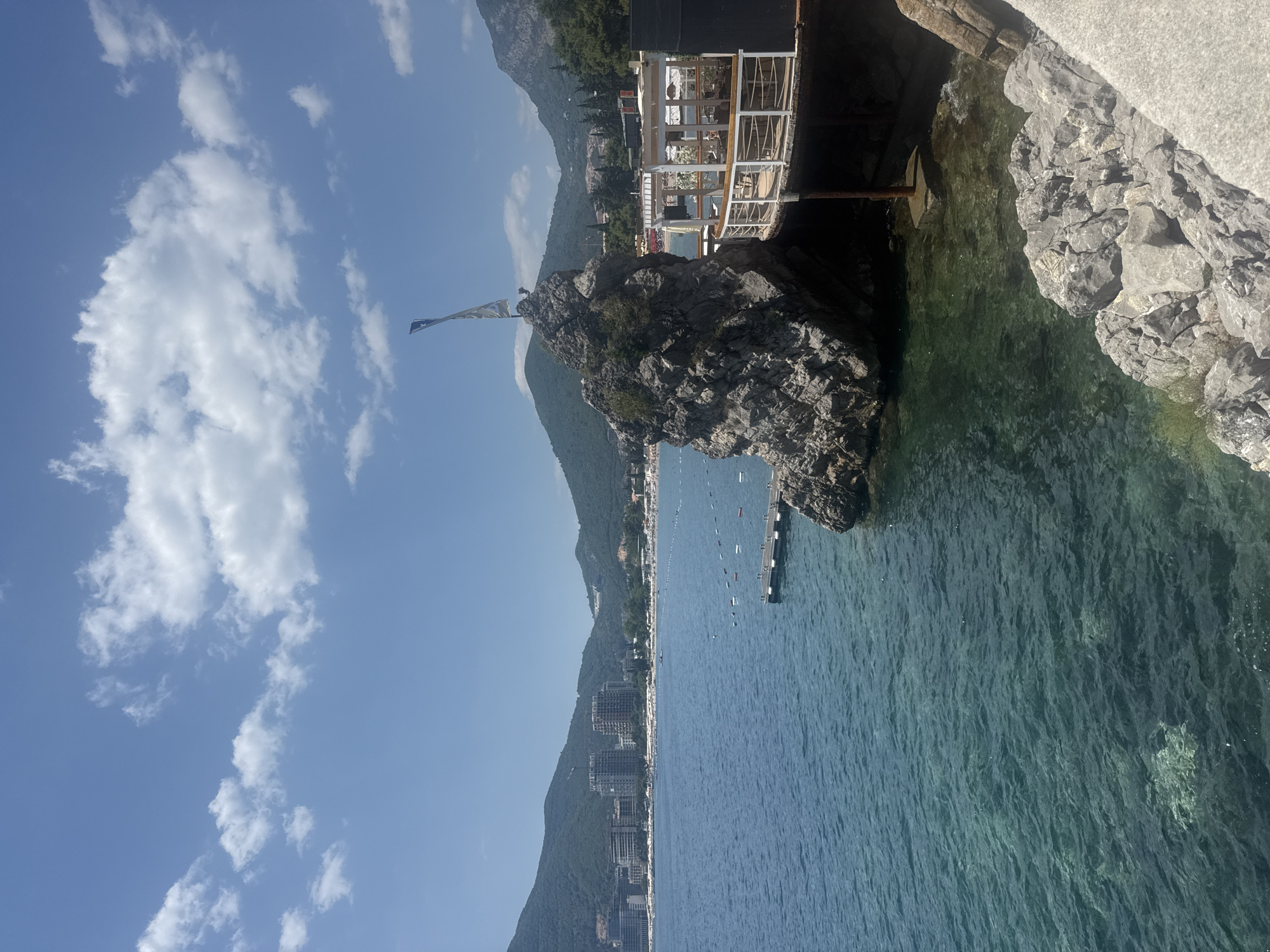 The image depicts a coastal scene, likely in Budva, Montenegro, on a clear, sunny day. The foreground features a rocky shoreline with the azure waters of the Adriatic Sea extending into the distance. A prominent, large rock formation juts out from the water near the shore, topped with a small patch of greenery and a flag that is not clearly discernible.
Behind the rock, a modern-looking structure with white railings and glass panels, possibly a restaurant or observation deck, is visible. It is built on stilts or a foundation extending over the water. The sea is calm, with small buoys and a distant pier dotting its surface.
Further in the background, a long stretch of coastline is lined with hotels and buildings, nestled against lush green mountains that form a dramatic backdrop. The sky above is a vibrant blue, punctuated by scattered white, fluffy clouds. The overall impression is one of a serene and picturesque Mediterranean summer day, with a mix of natural beauty and modern development. There is no visible text in the image.