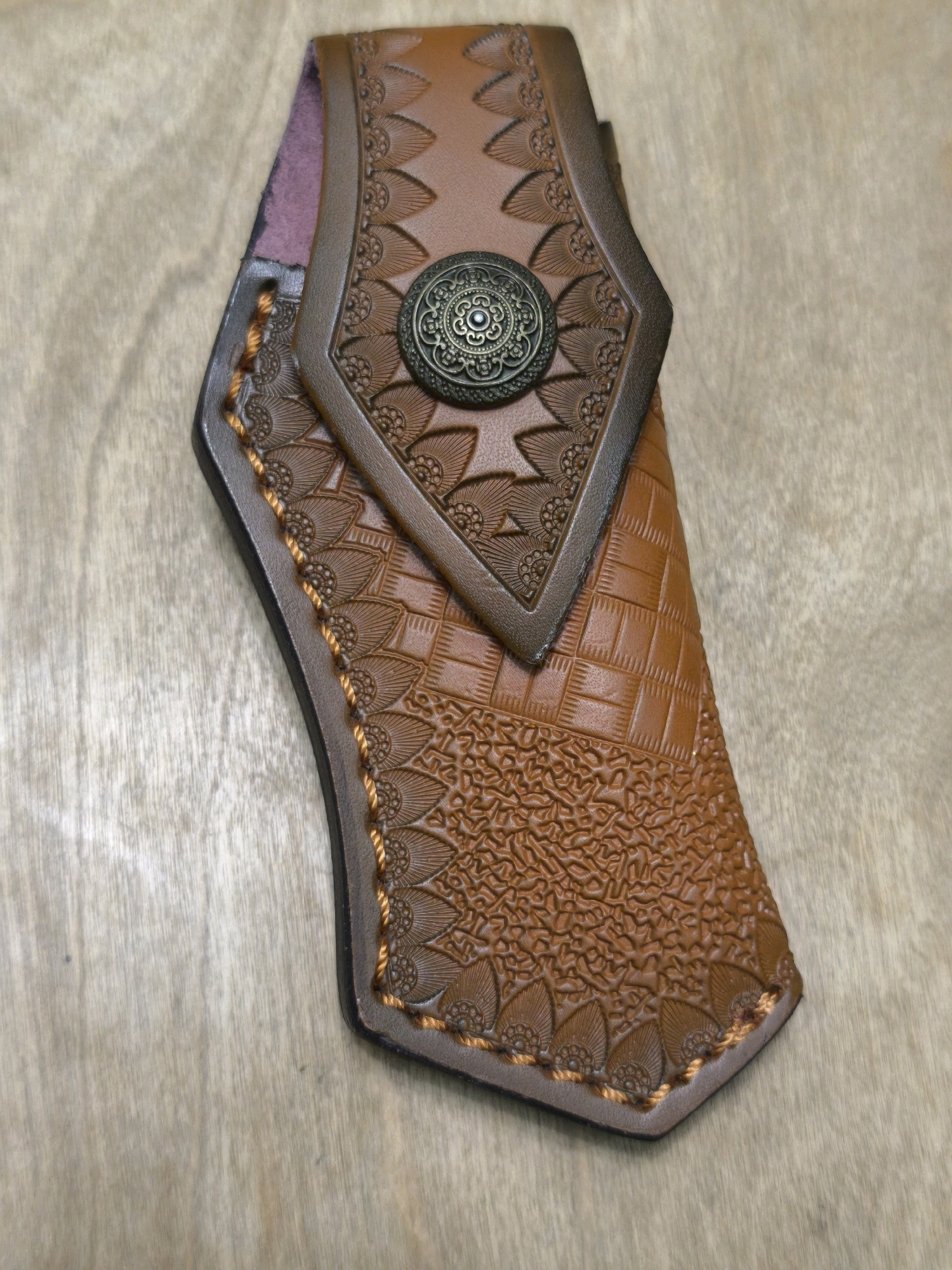 A brown leather sheath with decorative tooling and a metal embellishment lies on a wooden surface. The sheath features floral patterns and a woven texture. The wooden surface appears to be a table or countertop with a visible grain. The image is a top-down view.