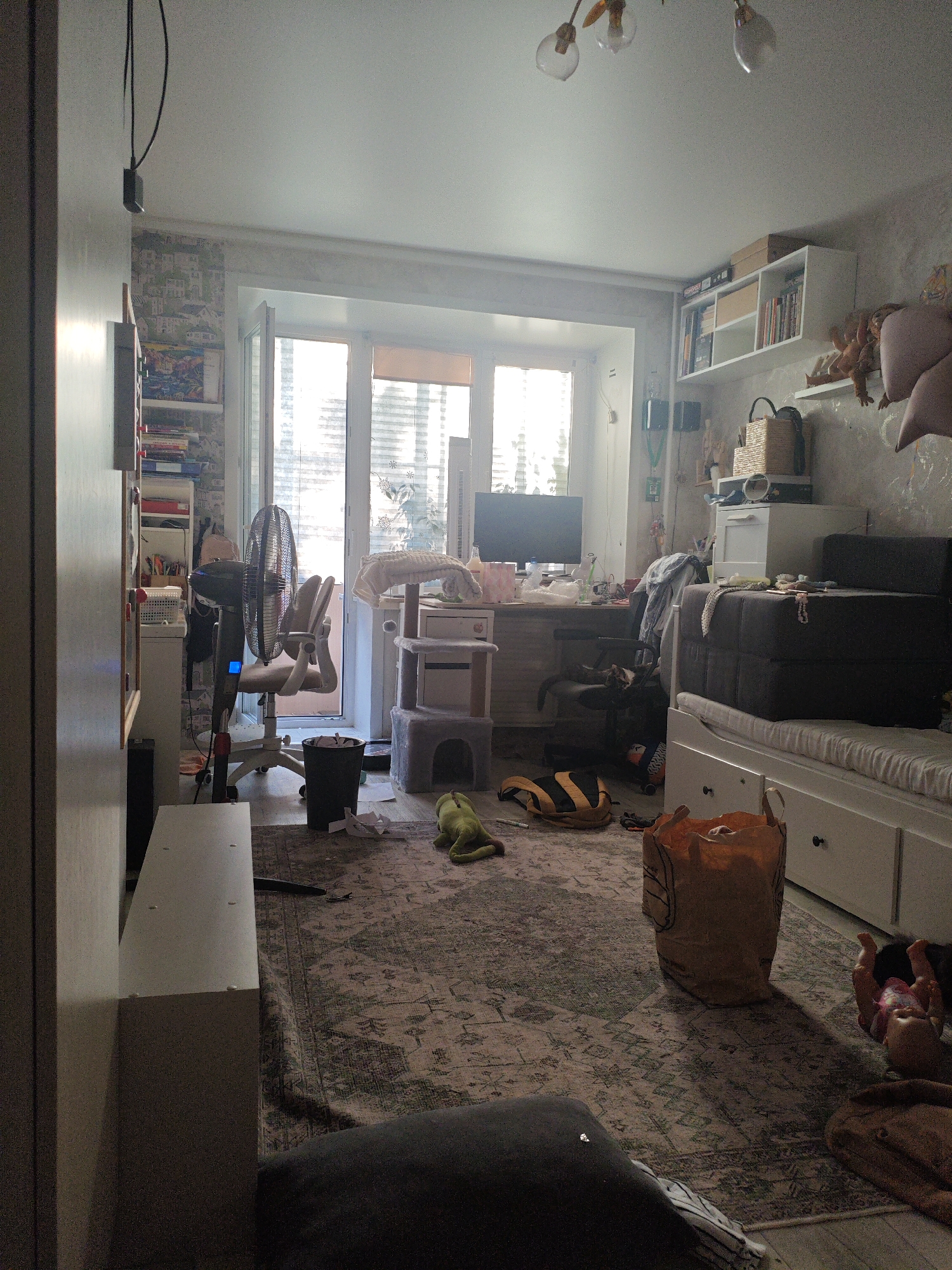This is a wide-angle, eye-level interior shot of a cluttered room that appears to be a child's bedroom or play area. The room is filled with various objects, suggesting a lived-in and somewhat unorganized space.
Dominant objects include a large fan in the center of the room, a desk with a computer monitor, a cat tree, a large sofa or daybed with drawers underneath, and several shelves filled with books and toys. There is also a trash can and a large brown paper bag on the floor, along with a green stuffed toy and a striped backpack. A doll is visible lying on the floor near the sofa.
The room is illuminated by natural light streaming in from a large window or balcony door that takes up a significant portion of the back wall. The blinds on the window are partially drawn, creating a bright but somewhat diffused light. The time of day appears to be daytime, given the strong natural light.
The overall impression is one of domestic disarray, with items scattered across the floor and surfaces. The wallpaper on the back wall features a pattern of houses. There are no people visibly present in the image. The location is identified as Berdsk municipality, Russia. No discernible text is visible in the image.
