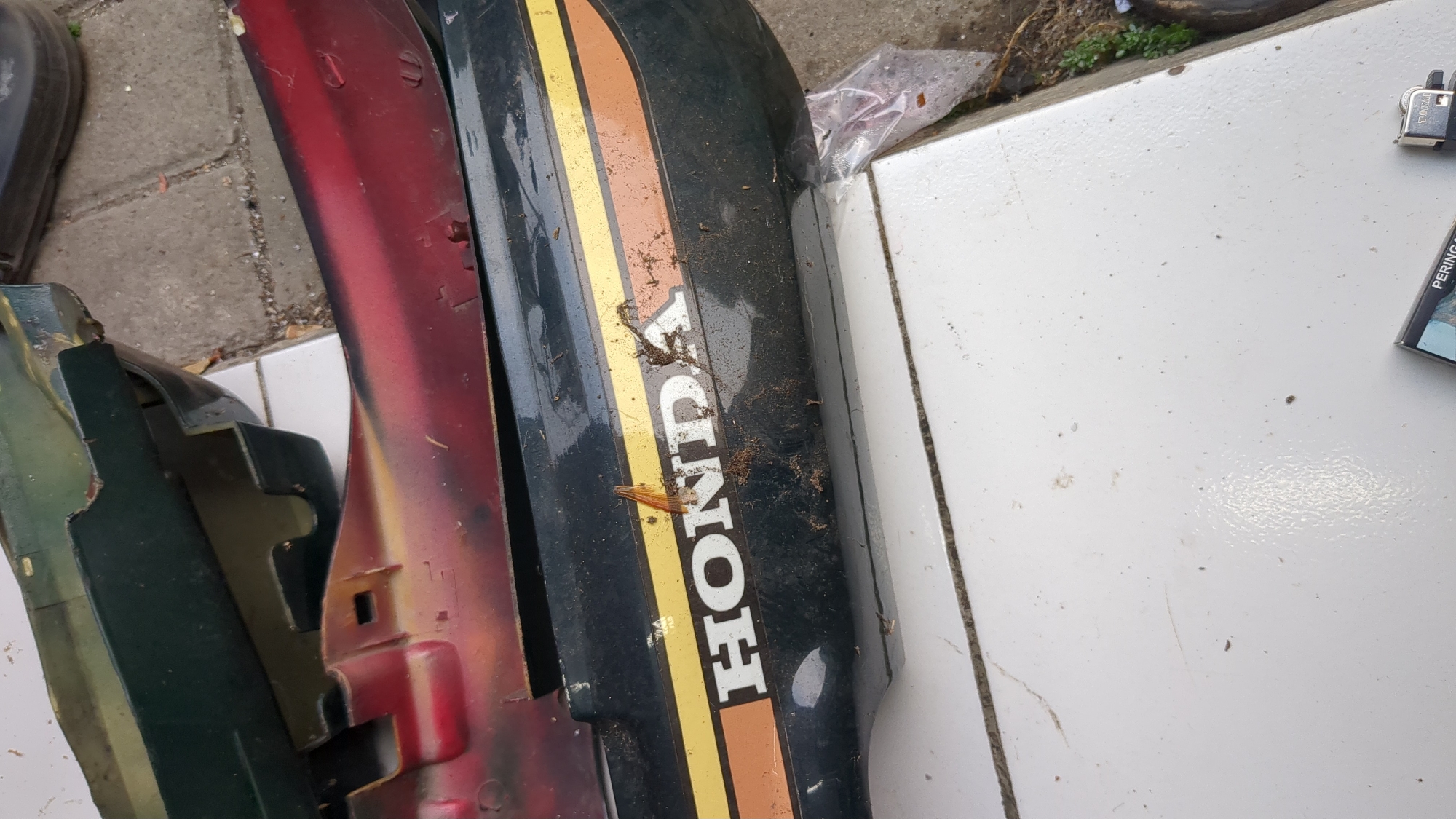 The image shows parts of a motorcycle, likely a fuel tank, with the "HONDA" logo clearly visible in white lettering on a black background, bordered by orange and yellow stripes. The motorcycle parts appear to be in disrepair, with some peeling paint on a red section above the black part. The objects are placed on a white tiled surface, with some dirt and debris scattered around. In the background, there is a paved area with some green vegetation. There are no people visible in the image, and no specific location in Purwakarta, Indonesia can be determined from the visual information provided.