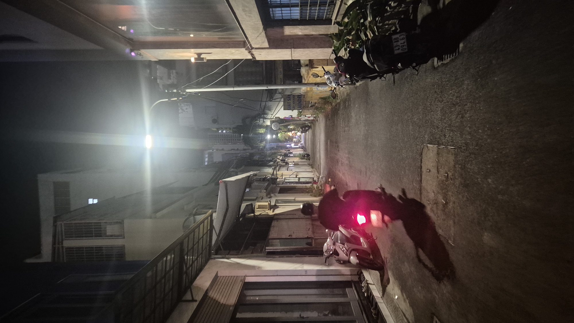 A nighttime, wide-angle, vertical shot captures a narrow alleyway in Đà Nẵng, Vietnam. The alley is flanked by buildings on both sides, with a single street lamp illuminating the scene from above, creating a bright lens flare. On the left, a light-colored building has a covered walkway with a metal grate roof extending over a sidewalk. Further down the alley on the left, a white motorcycle is parked next to a person on a dark-colored motorcycle, whose red brake light is illuminated. The person is wearing a dark shirt and a helmet. On the right side of the alley, a building has a textured facade and a dark-framed window with visible bars. A sign with Vietnamese text and the word "LAUNDRY" is visible on this building. Several motorcycles are parked along the right side of the alley, some with saddlebags. The asphalt alleyway has a wet sheen, reflecting the light. The overall atmosphere is urban and dimly lit.