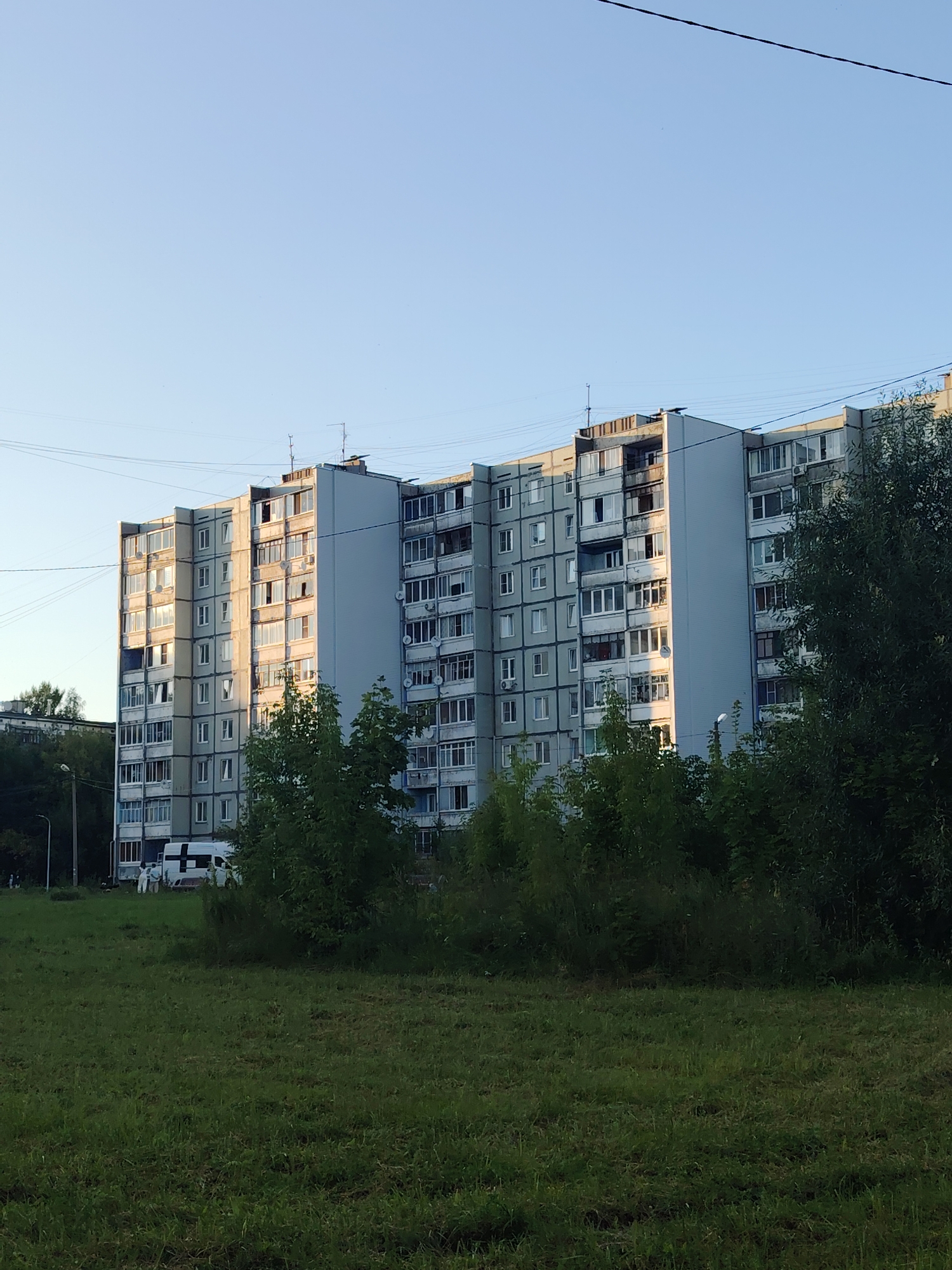 The image depicts a long, multi-story apartment building, characteristic of Soviet-era architecture, with a light gray facade and numerous windows. The building appears to be in the city of Konakovo, Russia, as suggested by the architecture and general environment. The building is situated behind a patch of green grass in the foreground and a line of dense green trees and bushes. The sky is clear and light blue, suggesting daytime, possibly late afternoon or early evening due to the warm light illuminating the facade of the building. Electrical wires are visible crisscrossing the upper portion of the image. On the left side of the building, a white van with a large black cross on its side, possibly an ambulance or medical transport, is parked near the entrance. Two figures, appearing to be wearing light-colored protective suits, are standing near the van and the building entrance. The scene appears calm and devoid of any overt signs of distress.