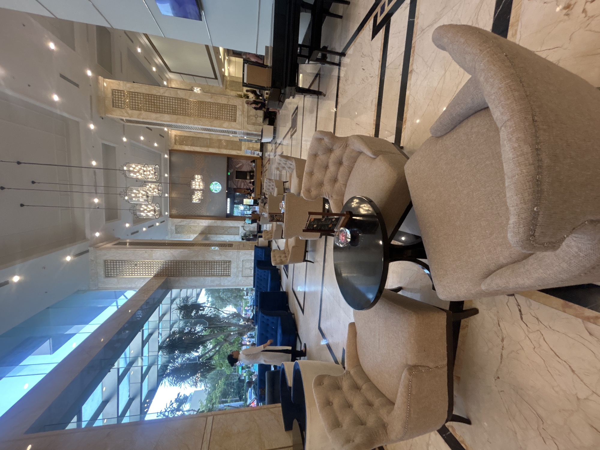 This image depicts the interior of a building, likely a hotel lobby, in Nha Trang, Vietnam. The scene is oriented lengthwise, showcasing a long reception area. The predominant colors are cream, beige, and white, creating a bright and airy atmosphere.
Foreground: Several plush armchairs upholstered in beige fabric are arranged around a small, round, black table. The armchairs are situated on a polished, marble-like floor with dark geometric patterns.
Midground: The reception area features a combination of seating arrangements, including blue couches and additional armchairs. A figure in a white uniform, possibly staff, is standing near the seating area, seemingly interacting with the space.
Background: The lobby extends into the distance, lit by chandeliers. The structure of the building is visible through large windows, revealing trees and sky outside. A piano is visible along the wall. The Starbucks logo is visible at the end of the lobby area.