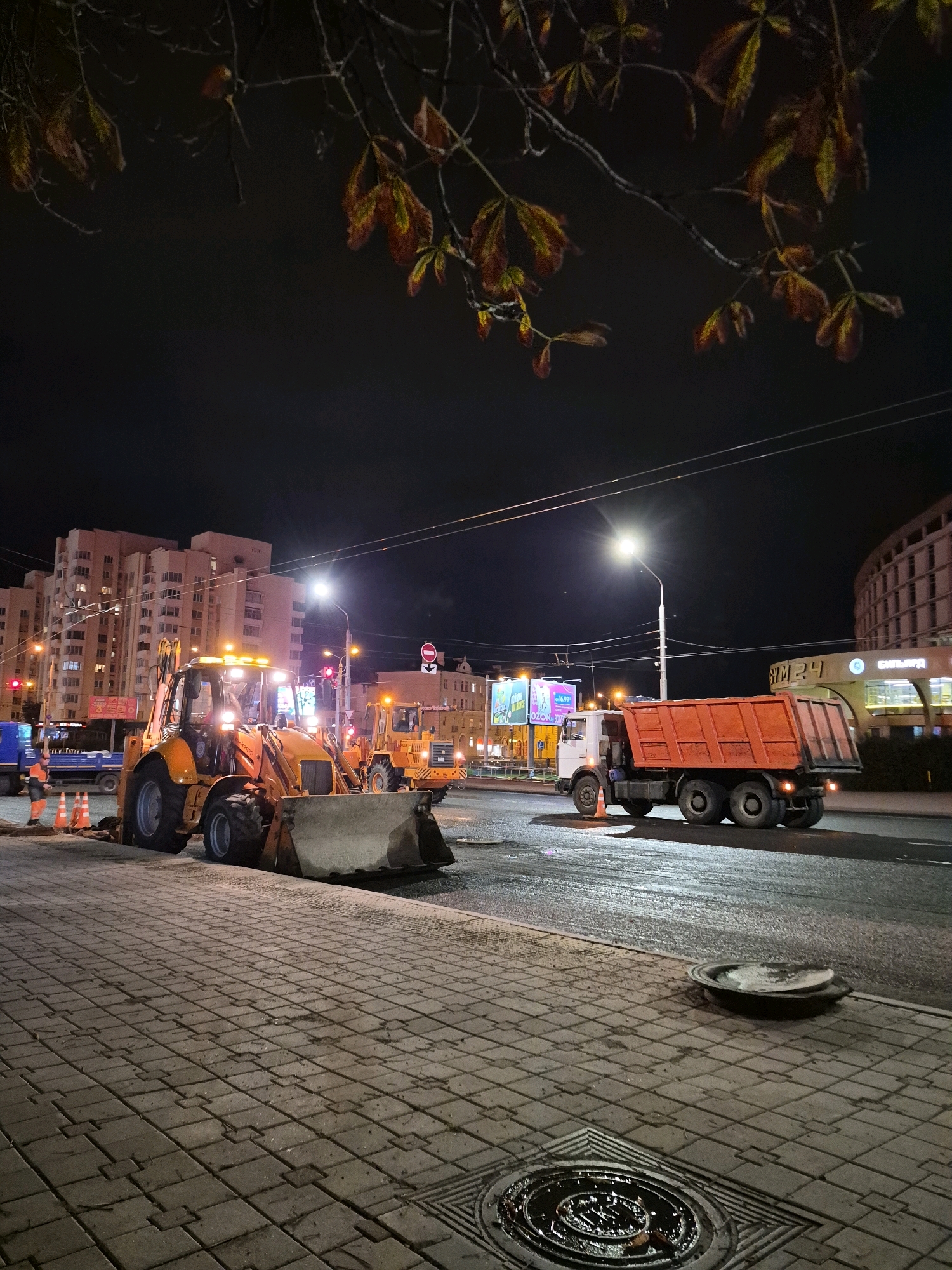An urban scene at night depicts road repair work on a wet asphalt surface. In the foreground, a cobblestone sidewalk is visible, with two manhole covers. A large orange backhoe loader is positioned on the road, its lights illuminated. Adjacent to it are several orange traffic cones and a blue truck. Further down the road, another yellow construction vehicle is present. An orange dump truck with its bed raised is also on the road. In the background, multi-story residential buildings with illuminated windows line the street, along with streetlights, traffic signals, and billboards. The image appears to be taken from a low angle, with tree branches and leaves overhead. The location is identified as Minsk, Belarus.