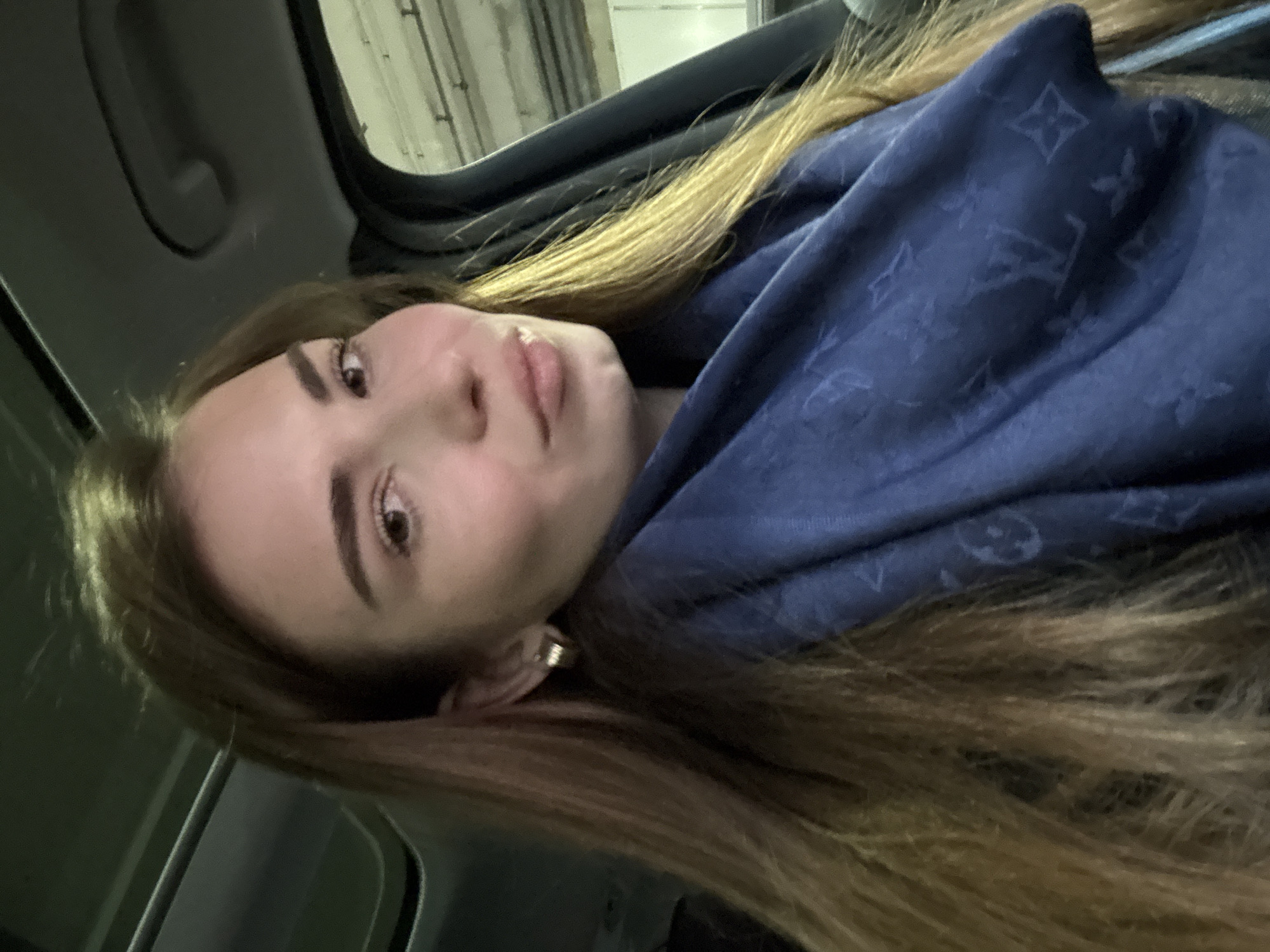 A woman is in a car, wearing a blue scarf with a pattern of Louis Vuitton logos. The woman is wearing an earring in her left ear. The car is moving and the background is blurry. The location cannot be confirmed to be in the city of Khimki, Russia.