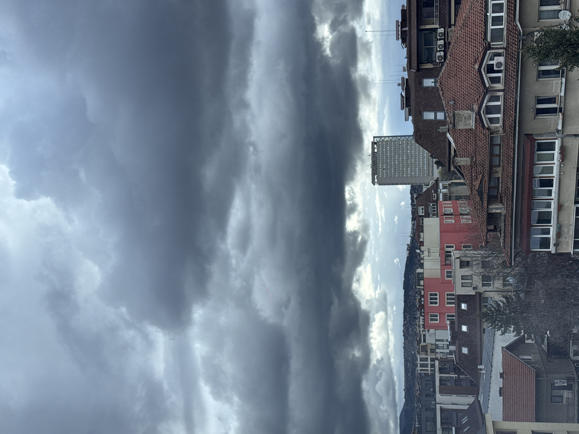 The image shows a cityscape under a heavily clouded sky. To the right are buildings of varying heights and styles. Some have red tile roofs, others are concrete structures, and some are painted in bright colors like red and white. Windows are visible across the facades.
The photograph appears to be taken in Sofia, Bulgaria, specifically in the area surrounding the "Hotel Rodina." This is recognizable from the architecture and layout of the buildings.