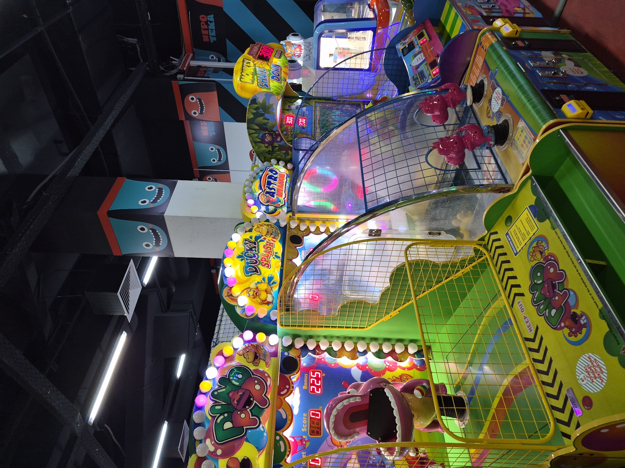 The image shows a lineup of brightly colored arcade games in an indoor space. The arcade games have names like "Ducky Splash," "Astro Invaders," and "Monkey Shake Down." The games feature lights and scores are displayed on digital displays. There are graphics on the walls in the background. A sign reads "ИГРО TEKA," which translates to "Game Room." The image likely originates in Tobolsk, Russia.
