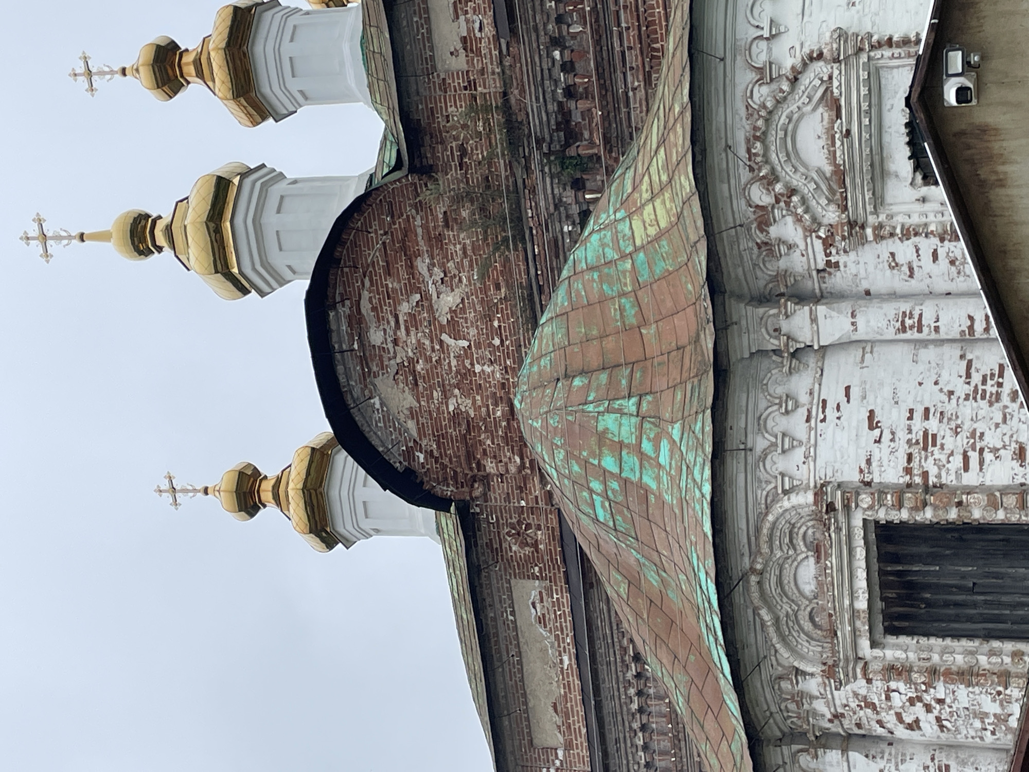 The image displays a section of a Russian Orthodox church in Kstynino, Russia. The foreground features a weathered, multi-faceted roof covered in oxidized copper, showing patches of green patina over a reddish-brown base. Behind this, the church's facade is visible, constructed from aged red brick with significant signs of deterioration, including crumbling mortar and exposed brickwork. Decorative elements, likely plasterwork, are present around arched windows and cornices, but these also show wear and tear, with some sections appearing damaged or missing. Above the brickwork, several gilded onion domes topped with crosses pierce the overcast sky. The domes are brightly polished and contrast sharply with the weathered architecture below. The overall impression is one of historical significance and enduring, yet deteriorating, beauty.