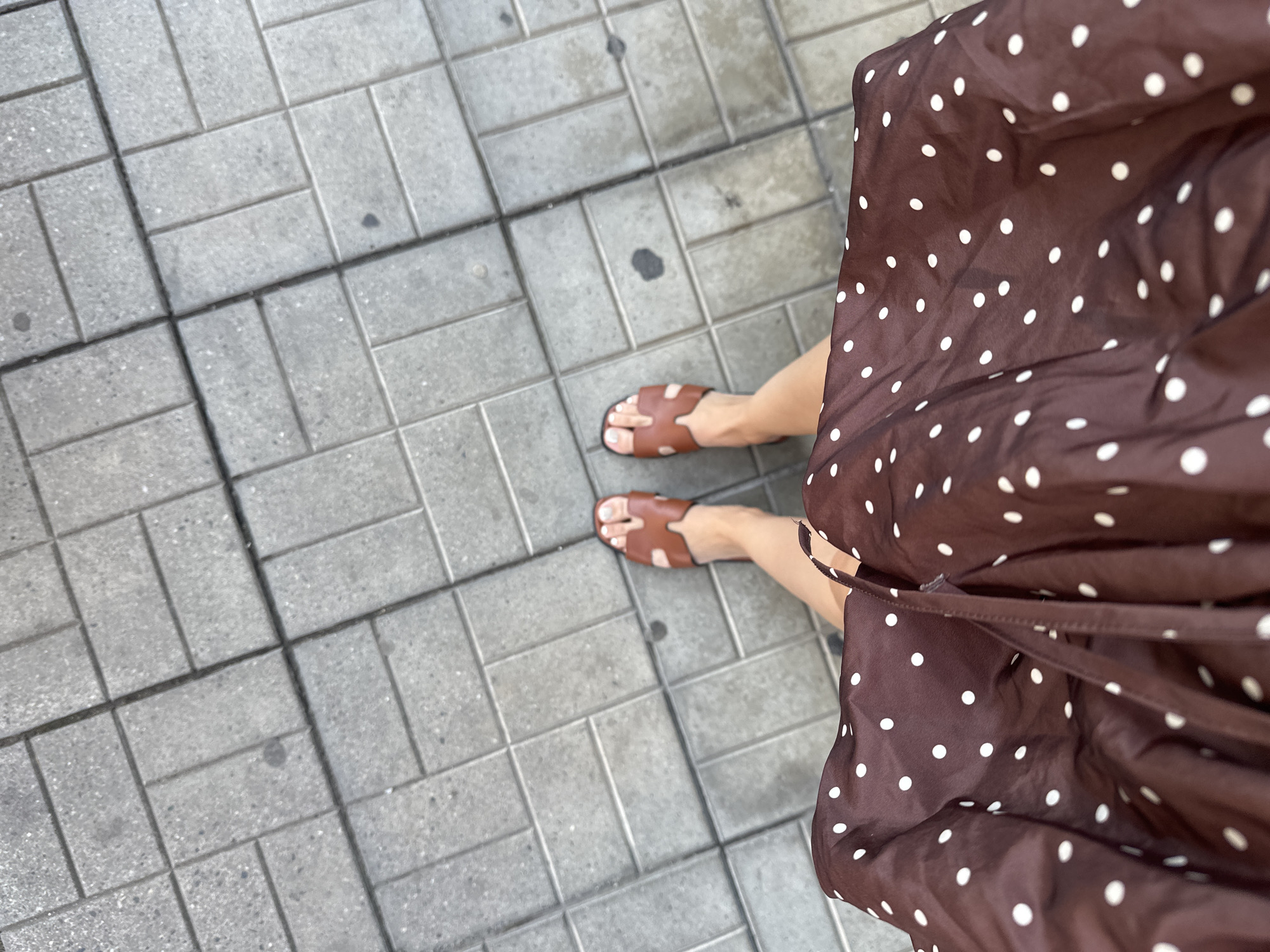 A high-angle shot shows a pair of legs walking on a sidewalk in Petrich, Bulgaria. The person is wearing brown slide sandals and a brown polka-dot skirt. The sidewalk is made of gray rectangular pavers.