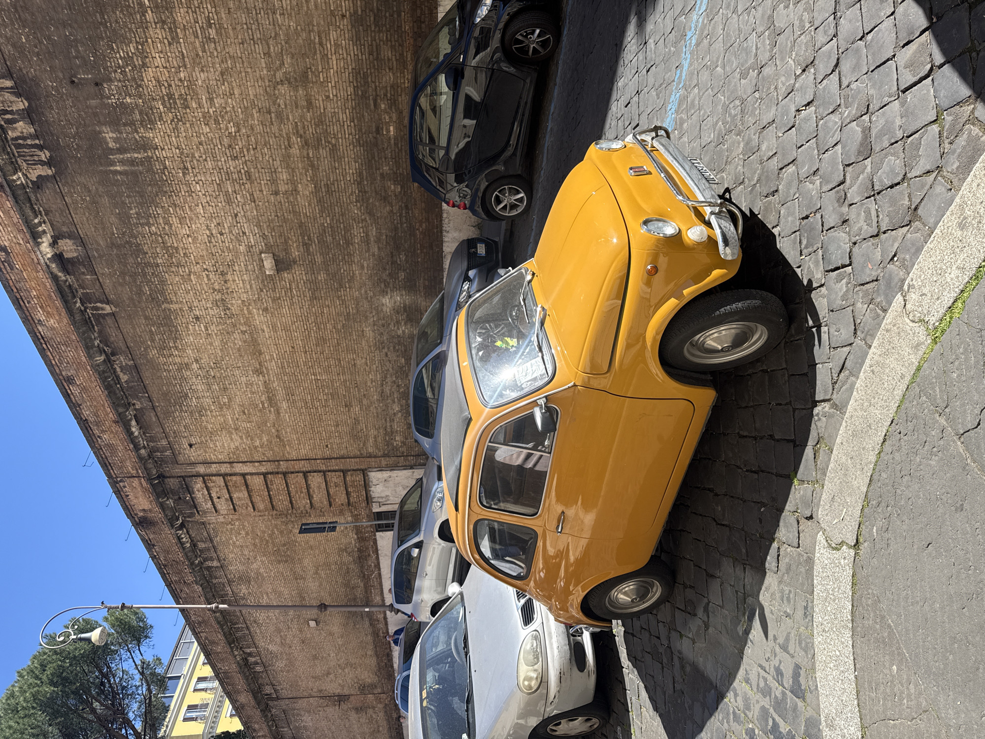 The image captures a street scene in Rome, Italy, lined with parked cars. The street is paved with cobblestones, and a tall brick building flanks one side of the road. The cars parked along the street include a distinctive yellow vintage vehicle at the front, followed by a Smart car and several other modern vehicles. A street lamp stands on the opposite side, near trees and buildings. The sky above is clear blue.