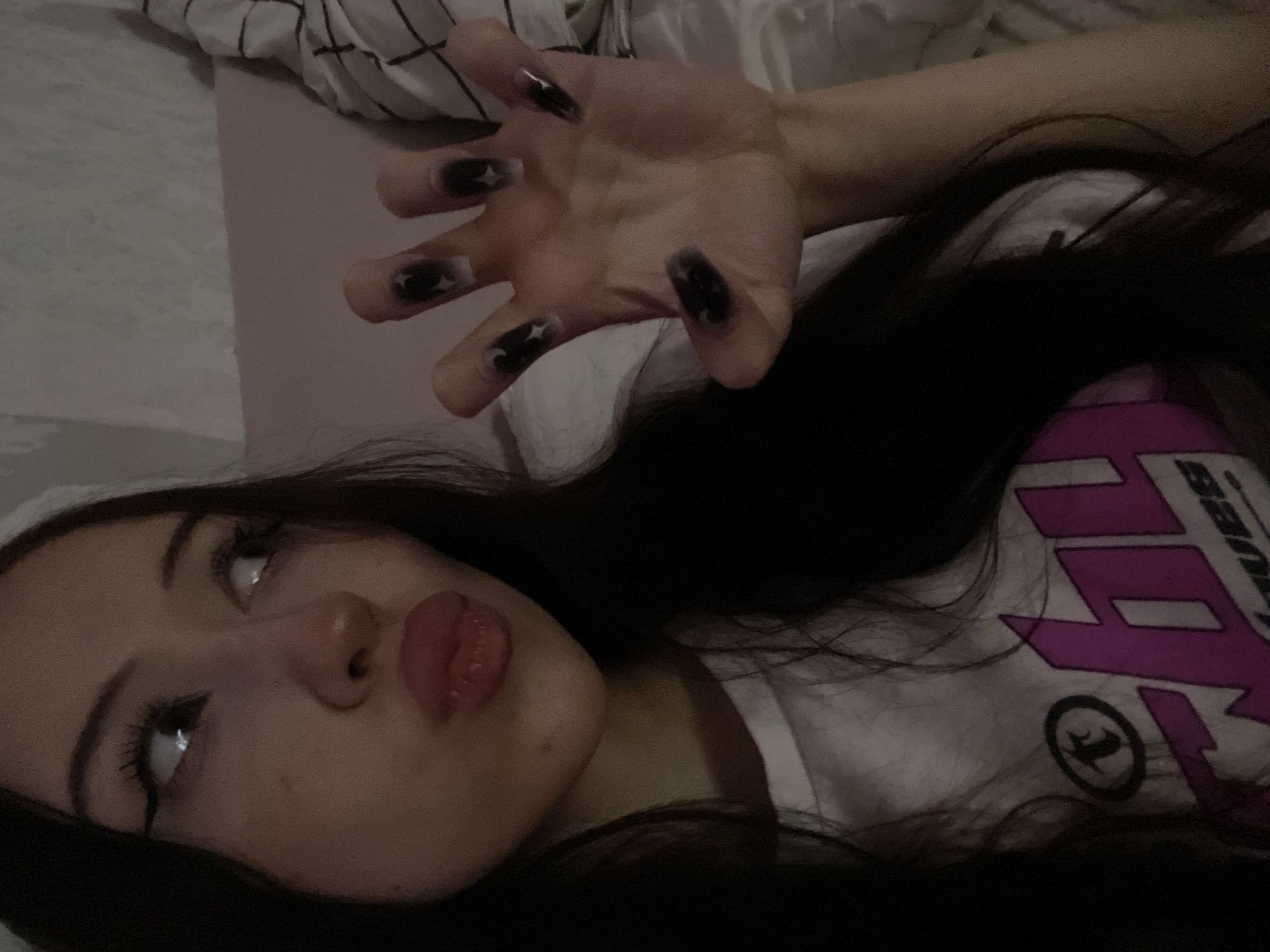The image shows a close-up of a young woman lying down, with her head tilted upwards. She has long, dark hair that partially obscures her face and chest. Her eyes are wide open, and she is wearing makeup, including winged eyeliner. Her lips are slightly parted, and she is wearing lipstick. Her right hand is raised in the foreground, with long, black fingernails, some of which have star designs. She is wearing a white t-shirt with a graphic design featuring pink lightning bolts and a circular logo. The background is blurred, but appears to be a bed with white and black striped bedding. The lighting suggests it is indoors, possibly in the evening or nighttime. There are no clear indicators of the weather or specific location within Votkinsk, Russia, other than the provided context. The overall mood is somewhat playful and attention-grabbing, due to the pose and makeup. The visible text on her shirt reads "BANDIT".