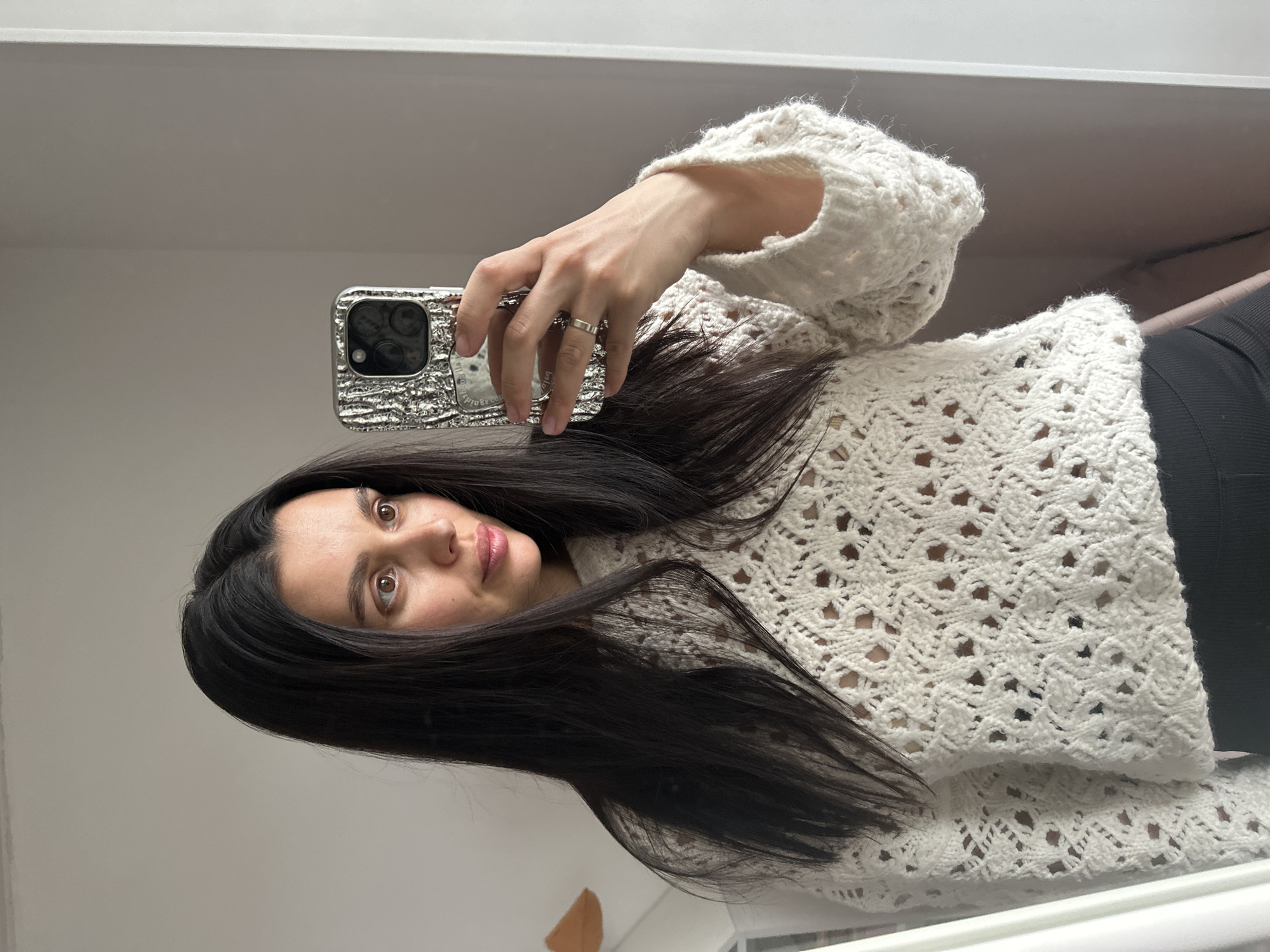 A woman with long, dark hair is taking a selfie in what appears to be an indoor setting. She is holding a phone in her right hand, which has a shiny silver case. She is wearing a loose, white knitted sweater with a pattern of holes. Part of a black garment, possibly pants or a skirt, is visible at the lower right. The background is a plain, light-colored wall. The image does not provide information to determine if the photo was taken in Chaikovsky, Russia.
