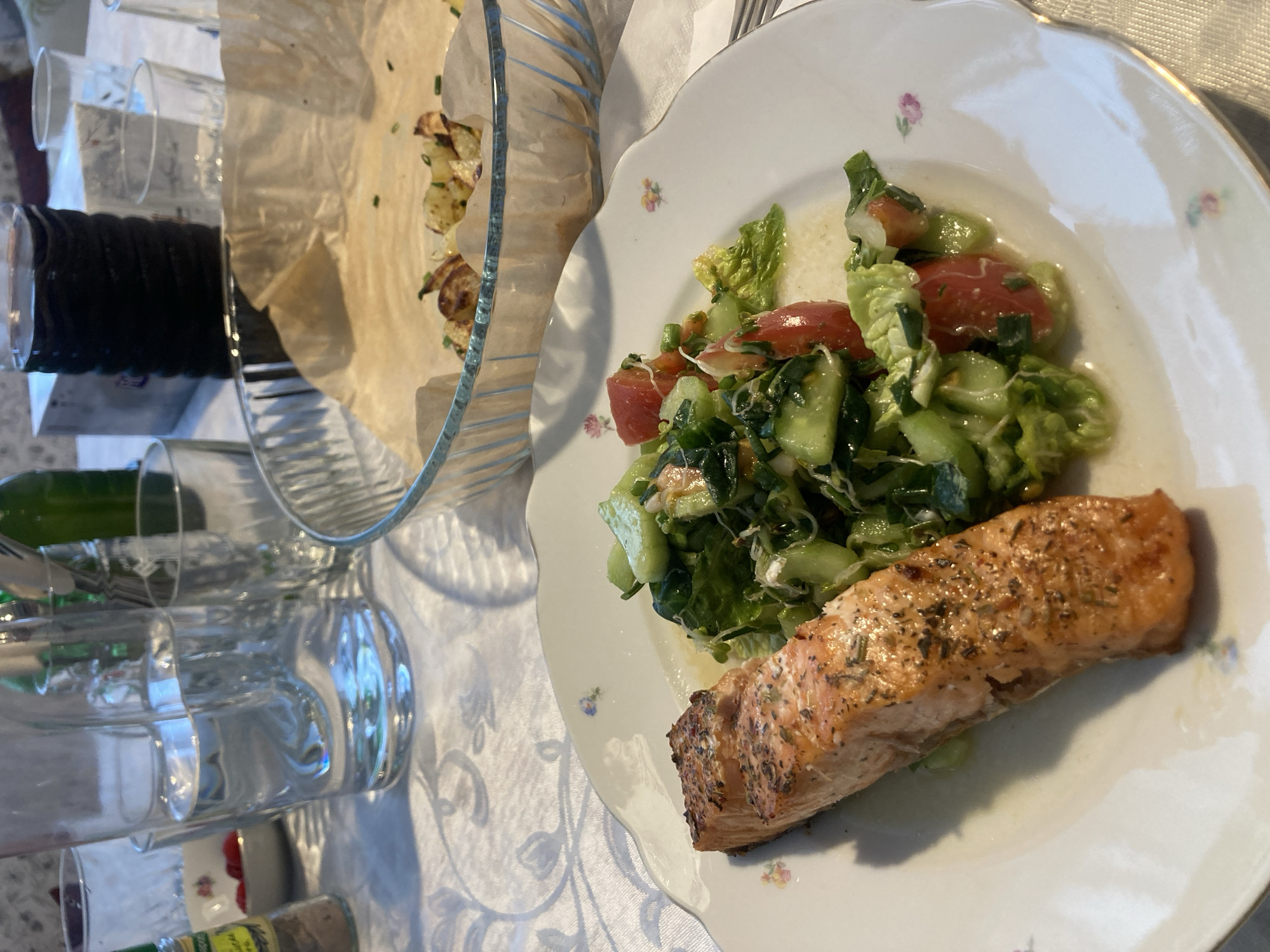 The image shows a plate of food with a piece of salmon and a salad with tomatoes and cucumbers. Next to the plate is a glass bowl with some cooked potatoes. There are several glasses on the table. The setting appears to be a dining table, possibly in a home, covered with a white tablecloth. The location is unknown.