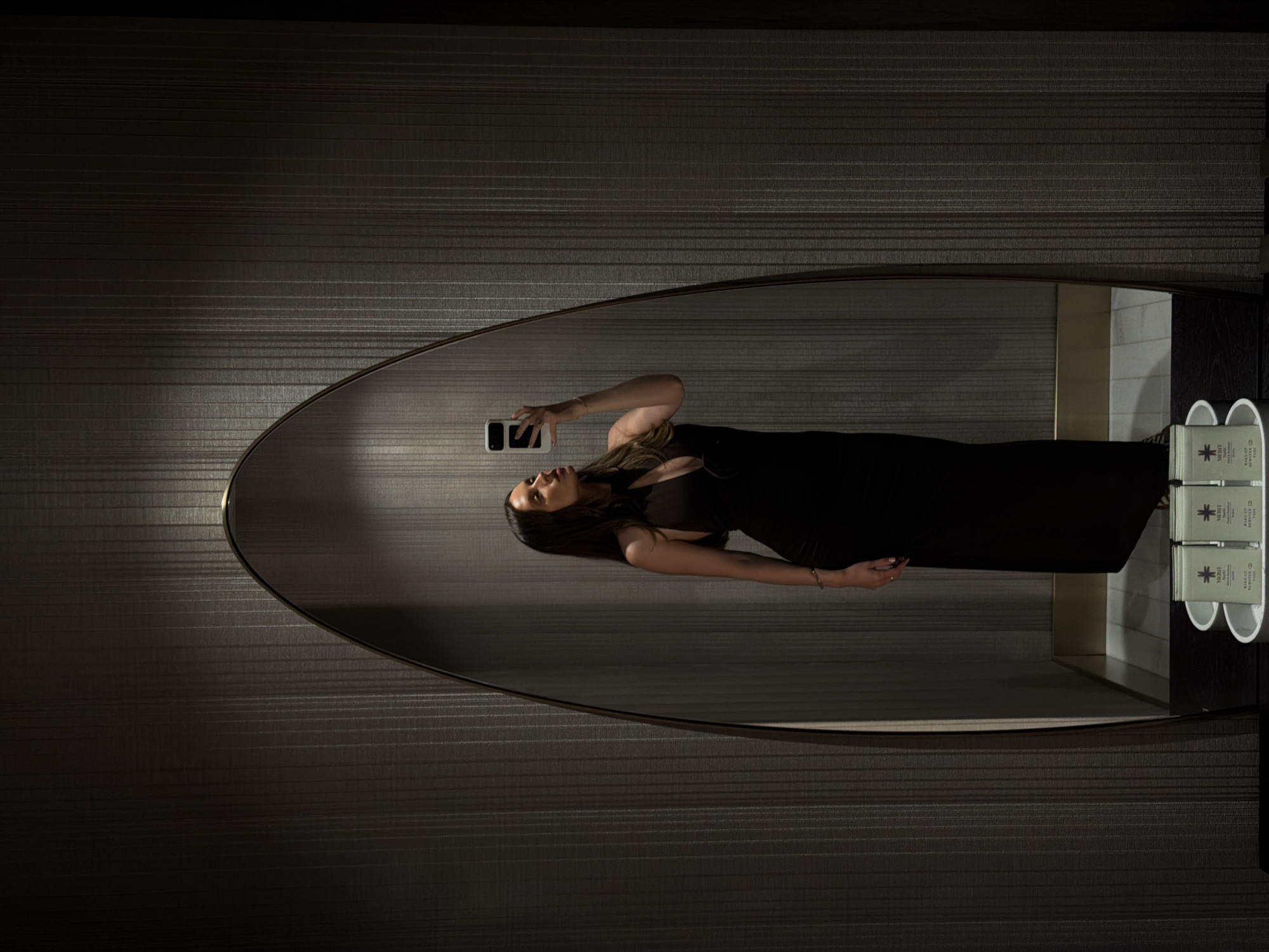 An interior shot captures a woman taking a mirror selfie in a dimly lit, upscale room within a private residence or hotel in Budva, Montenegro. The central feature is a tall, oval-shaped mirror with a thin dark frame, reflecting the woman and a portion of the room. The woman, with long dark hair, wears a form-fitting, sleeveless black gown with a deep V-neckline. She holds a white-cased smartphone in her right hand, angled to capture her reflection. Her left arm is slightly bent, revealing a simple bracelet. The room's walls are covered in dark, vertically textured wallpaper. Overhead lighting casts a bright spot onto the upper portion of the mirror and the wall above it, creating a contrast with the otherwise subdued illumination. A dark shelf beneath the mirror holds a white tray containing at least four small, rectangular packages labeled "MERIDT" and "WAKE-UP EYE REPAIR PADS." The reflection in the lower part of the mirror shows a light-colored floor, possibly tiled.
