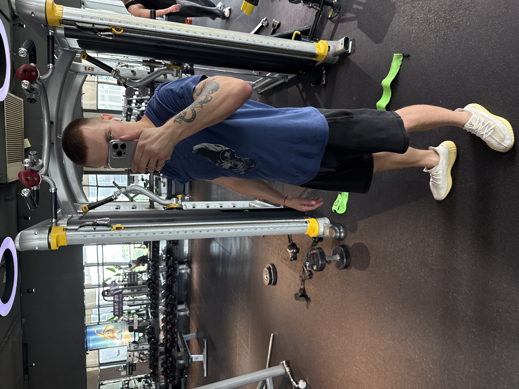 A young adult male with a buzz cut is taking a selfie in a gym located in the Haizhu District, China. He is wearing a navy blue t-shirt with a skull graphic and black shorts. He has white wireless earbuds in his ears and tattoos on his right arm. In the background, various gym equipment is visible, including weight racks filled with dumbbells, weight machines, and benches. The floor of the gym is dark rubber. A bright green resistance band is lying on the floor near his feet. Another person, wearing a black tank top and grey pants, is partially visible to the right, walking away from the camera. The gym appears to be well-lit with overhead lighting and natural light coming from large windows in the background, which offer a view of urban buildings.