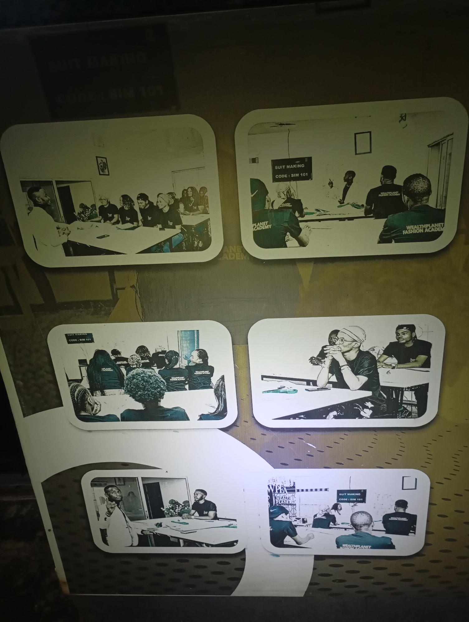 The image displays a collage of several black and white photographs, possibly mounted on a wall in an indoor setting. The photographs depict scenes related to fashion or tailoring education.
In the top left photograph, a man in a white jacket appears to be lecturing or instructing a group of people seated at a table. The people are wearing dark t-shirts, some with visible text.
The top right photograph shows a similar scene with a group of individuals gathered around tables. A sign on the wall reads "SUIT MAKING CODE: SIM 101". The individuals are also wearing dark t-shirts with visible text.
The middle left photograph features a group of individuals from behind, seated at tables in what appears to be a classroom or workshop. They are wearing dark t-shirts with the text "WEALTHPLANET FASHION ACADEMY".
The middle right photograph shows three individuals seated at a table. One person in the foreground is wearing glasses and a headscarf, looking thoughtful. The other two are wearing t-shirts.
The bottom photograph is a smaller image showing a man in a suit gesturing while speaking to another man seated at a table. There is a floral arrangement on the table.
A second smaller photograph at the bottom right shows a group of people in a room, with one person standing and addressing the seated individuals. The sign "SUIT MAKING CODE: SIM 101" is visible on the wall.
The background of the collage appears to be a textured surface, possibly wallpaper or a wall treatment, with a repeating pattern. The lighting in the overall image is uneven, suggesting it might have been taken in low light conditions or with direct flash. There are no discernible architectural features or landmarks that would indicate a specific city or location.