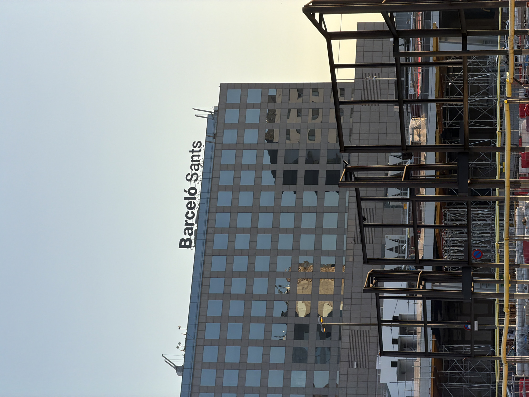 The image shows a tall, modern building with a facade of glass windows. The text "Barceló Sants" is visible vertically on the left side of the building, indicating it's likely a hotel. The building is surrounded by construction, with scaffolding, metal beams, and yellow safety barriers visible in the foreground. The construction suggests an ongoing project or renovation. The sky is bright and clear, suggesting it is daytime, possibly late afternoon given the warm tones in the sky and reflections. The reflection in the glass windows of the building shows other buildings and the construction site, adding depth to the scene. The overall impression is one of urban development and activity in Barcelona, Spain.
