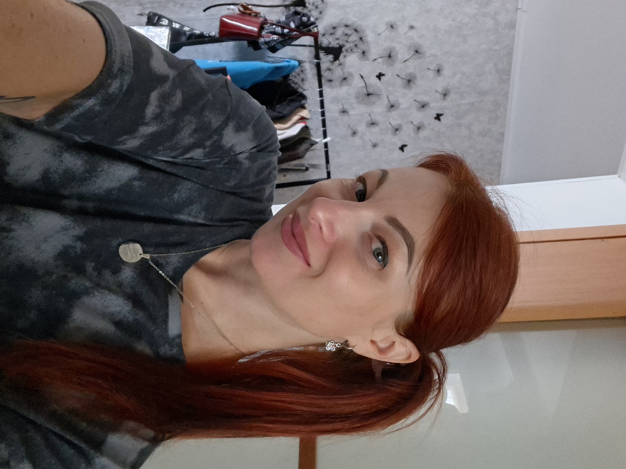 A woman with red hair pulled into a ponytail, wearing a dark tie-dye t-shirt and a silver necklace with a circular pendant, is taking a selfie. She is wearing a silver earring with a small flower and a feather-like charm. In the background, there is a clothing rack with several garments hanging on it, a red purse, and a wall decorated with dandelion and butterfly decals. The environment appears to be indoors, possibly a room in a home in Myadzyel, Belarus.