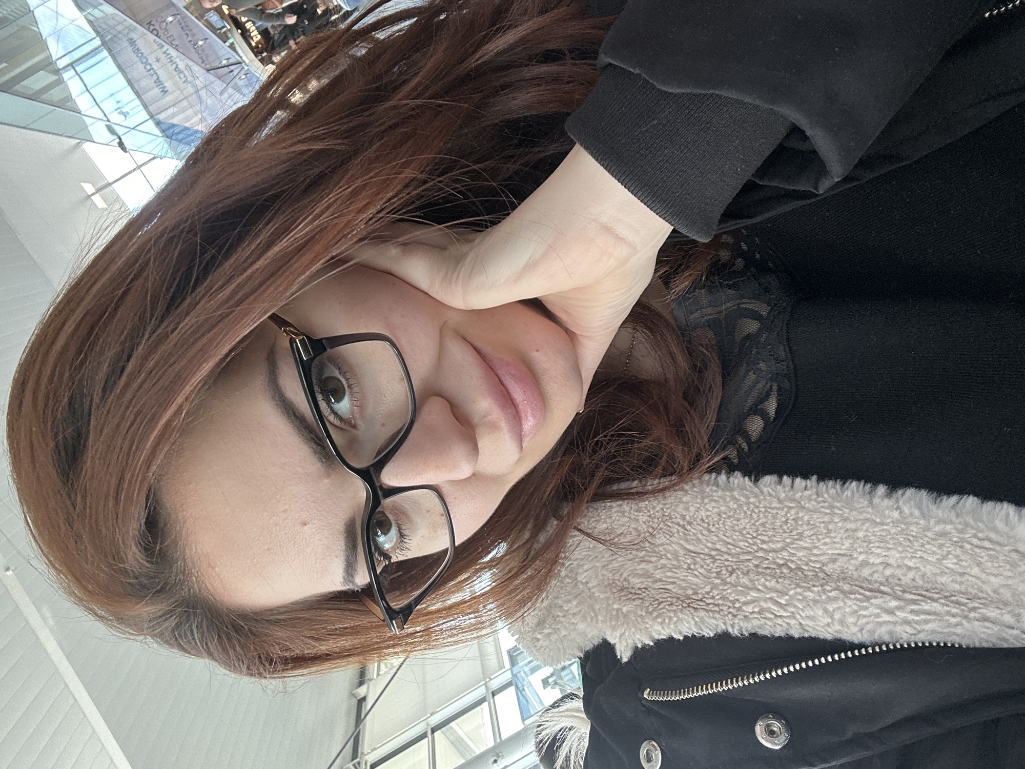 A woman with auburn hair and glasses is positioned close to the camera. She has fair skin and is wearing a black coat with a light-colored furry lining. The woman's left hand is placed against her cheek.
Reflections of the surroundings are visible in the background, indicating an interior space with large windows. The reflections include signs that suggest the location is Wrocław Airport (WRO), in Wrocław, Poland. There are people visible in the background, suggesting that this could be a public waiting area.