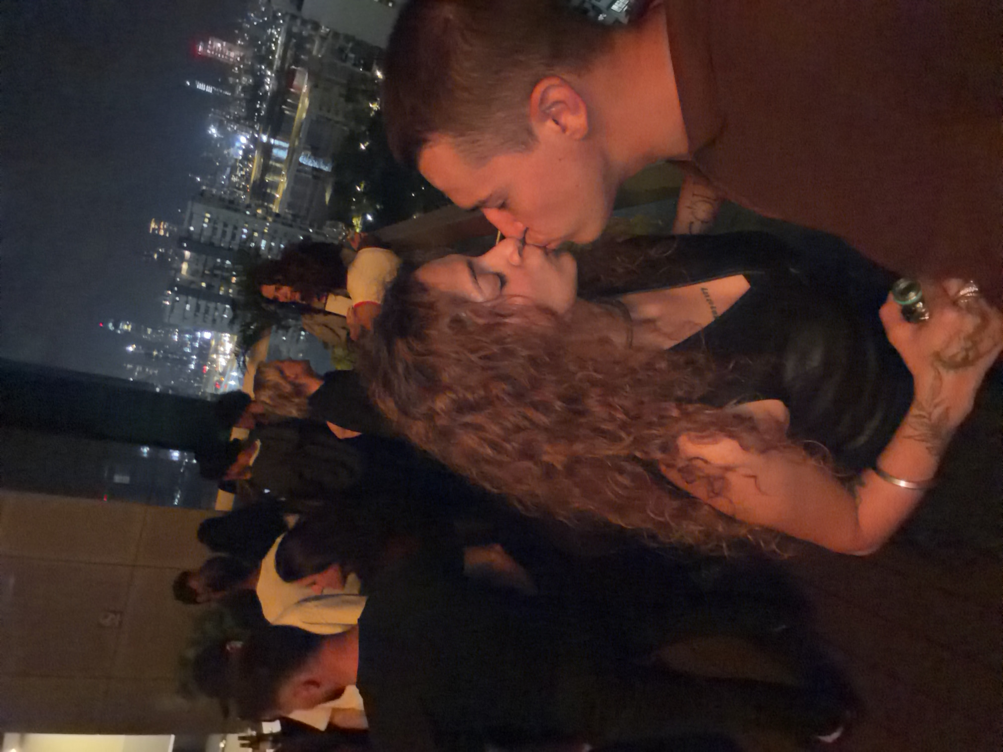 The image depicts a couple kissing on a rooftop overlooking a city skyline at night. The man on the right is wearing a dark brown t-shirt. The woman on the left has long, curly brown hair, a black top, and is wearing a silver bracelet on her left arm. She also has tattoos visible on her arm. In the background, other people are visible in the dimly lit area, some standing and some sitting. Beyond the rooftop, the city of Mumbai is illuminated, with numerous buildings, lights, and a body of water. The overall atmosphere suggests a social gathering in a vibrant urban environment.