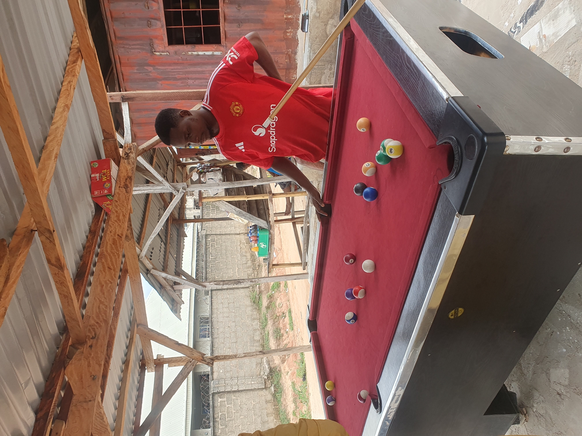A man wearing a red Manchester United jersey is playing pool at an outdoor establishment in Lokoja, Nigeria. The pool table has a red felt surface and is covered with several billiard balls, including a cue ball. The man is holding a cue stick and appears to be in the process of taking a shot. The background shows a rustic, open-air structure with a corrugated metal roof and wooden beams. A brick wall and some buildings are visible in the distance. The scene suggests a casual recreational setting.