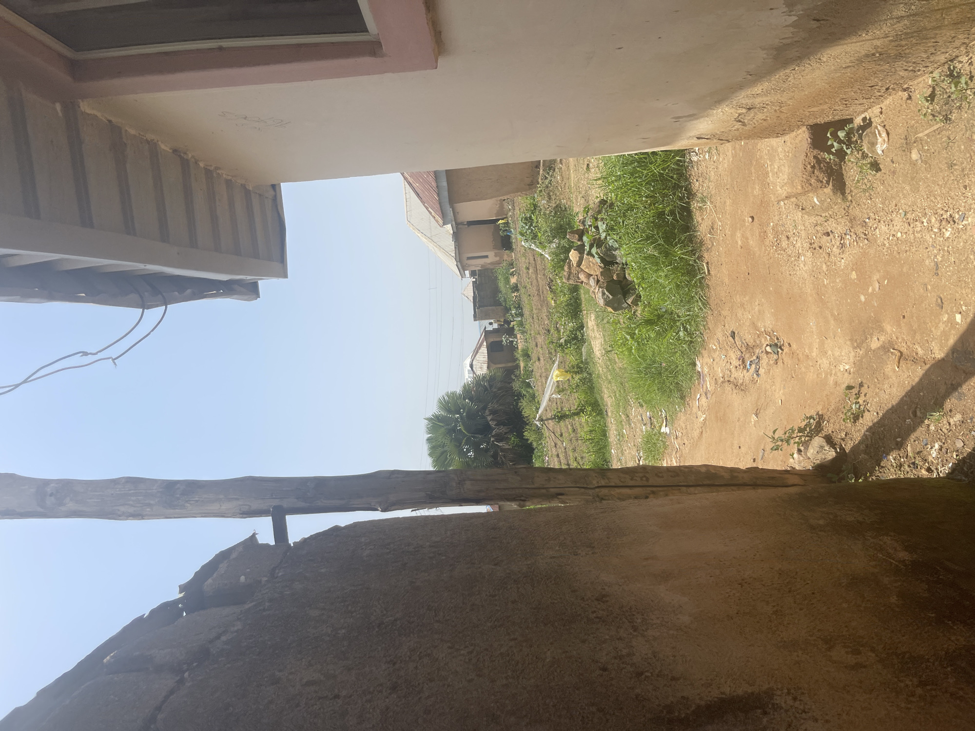 This media file shows an outdoor scene in Rafin Par, Nigeria, likely during the daytime under clear skies. The perspective is from a narrow passage between two structures, looking out towards a residential area.
On the left side of the frame, a portion of a building's exterior is visible, featuring a light-colored wall with some horizontal grooved detailing underneath. A small section of a window frame with a pinkish trim is also present. Wires are seen hanging down from this structure.
The right side of the frame shows a dirt path with scattered rocks and some sparse green vegetation. Further into the background, several small, simple houses are visible, some with corrugated metal roofs. There's an area with longer, thicker green grass, and a distinct palm tree stands out amongst the trees and buildings. The ground appears dry and dusty, typical of an arid or semi-arid environment. The overall scene suggests a rural or semi-urban residential setting.
There are no people visible in the image, and no specific activity or event is clearly taking place. The lighting indicates it is daytime, and the clear sky suggests fair weather. The presence of simple housing and the unpaved paths are strong location cues for the Nigerian setting. No visible text is present in the image.