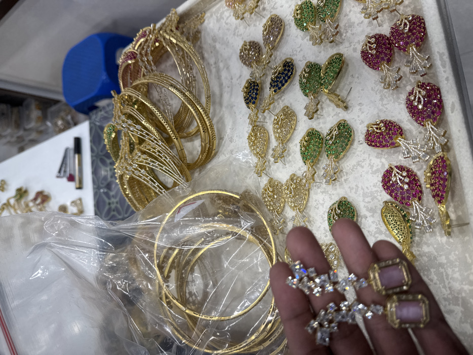 The image showcases a collection of glittering jewelry. Gold bangles, some adorned with sparkling stones, are clustered together. Earrings featuring colorful stones and intricate designs lie on a white surface, adding vibrancy to the scene. A hand presents a pair of earrings, one a cluster of white stones and the other a pink rectangular stone, highlighting the craftsmanship and elegance of the jewelry collection.