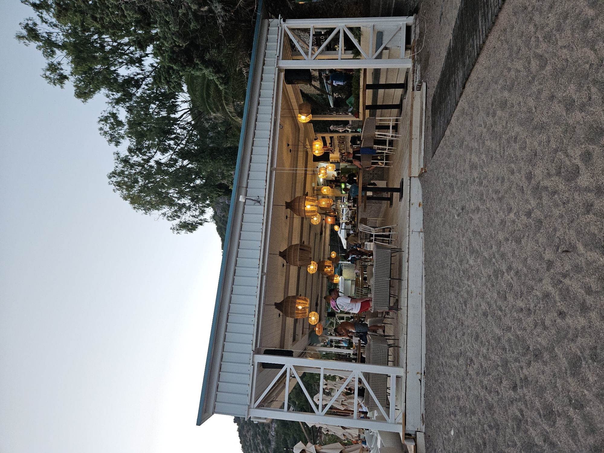 The image depicts an outdoor beachside establishment in Kamenovo, Montenegro, at dusk. The structure is a covered open-air bar and seating area with a white metal frame and a light-colored corrugated roof. Woven pendant lights, emitting a warm glow, are suspended from the ceiling, illuminating the interior and the surrounding tables and chairs. The seating area includes several tables with chairs and benches, some of which are occupied by patrons. A bar counter is visible inside, with bottles and glasses on display. In the foreground, dark sand covers the beach, with a wooden walkway leading to the establishment on the right. Lush green trees and a hillside are visible in the background, under a pale, fading sky. Several people are present, some standing and conversing, others seated. One person in the foreground wears a white t-shirt and red shorts, while another, closer to the bar, is dressed in a white t-shirt and blue shorts. A person wearing a red and white headscarf is also visible near the entrance. The overall scene suggests a relaxed atmosphere at a seaside resort.